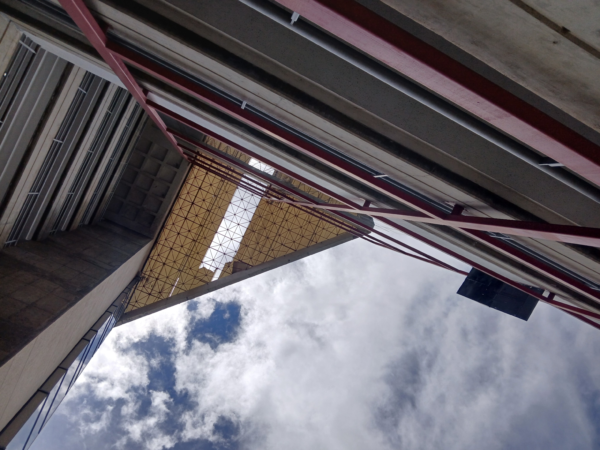 The image shows an upward perspective of building structures against a cloudy sky. Concrete beams and red metal supports frame the view. A triangular section with a crisscross pattern lattice provides partial coverage, while a dark rectangular element is affixed to one of the beams. The location is Ciudad Guayana, Venezuela, as it shows details of the architecture and infrastructure in that city.