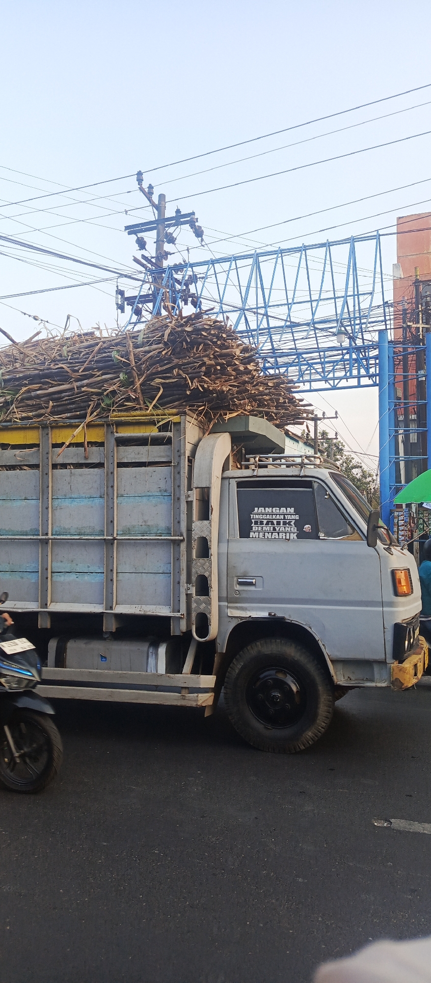 A light gray truck loaded with sugarcane stalks is driving on a road in Malang, Indonesia. The truck is a typical Indonesian " Colt Diesel" truck. The sugarcane is piled high on the truck's bed, extending above the cab. The truck has a license plate that appears to be an Indonesian license plate. A motorcycle is visible on the left side of the truck, partially obscuring the truck's tires. The truck has a sticker on its window that says "JANGAN TINGGALKAN YANG BAIK DEMI YANG MENARIK" which translates to "Don't leave the good for the attractive." The sky is bright and overcast. In the background, there are power lines, a blue metal structure that could be a bridge or an overpass, and buildings. A green tarp is visible on the right side of the image. The road surface is asphalt and appears to be a busy street.