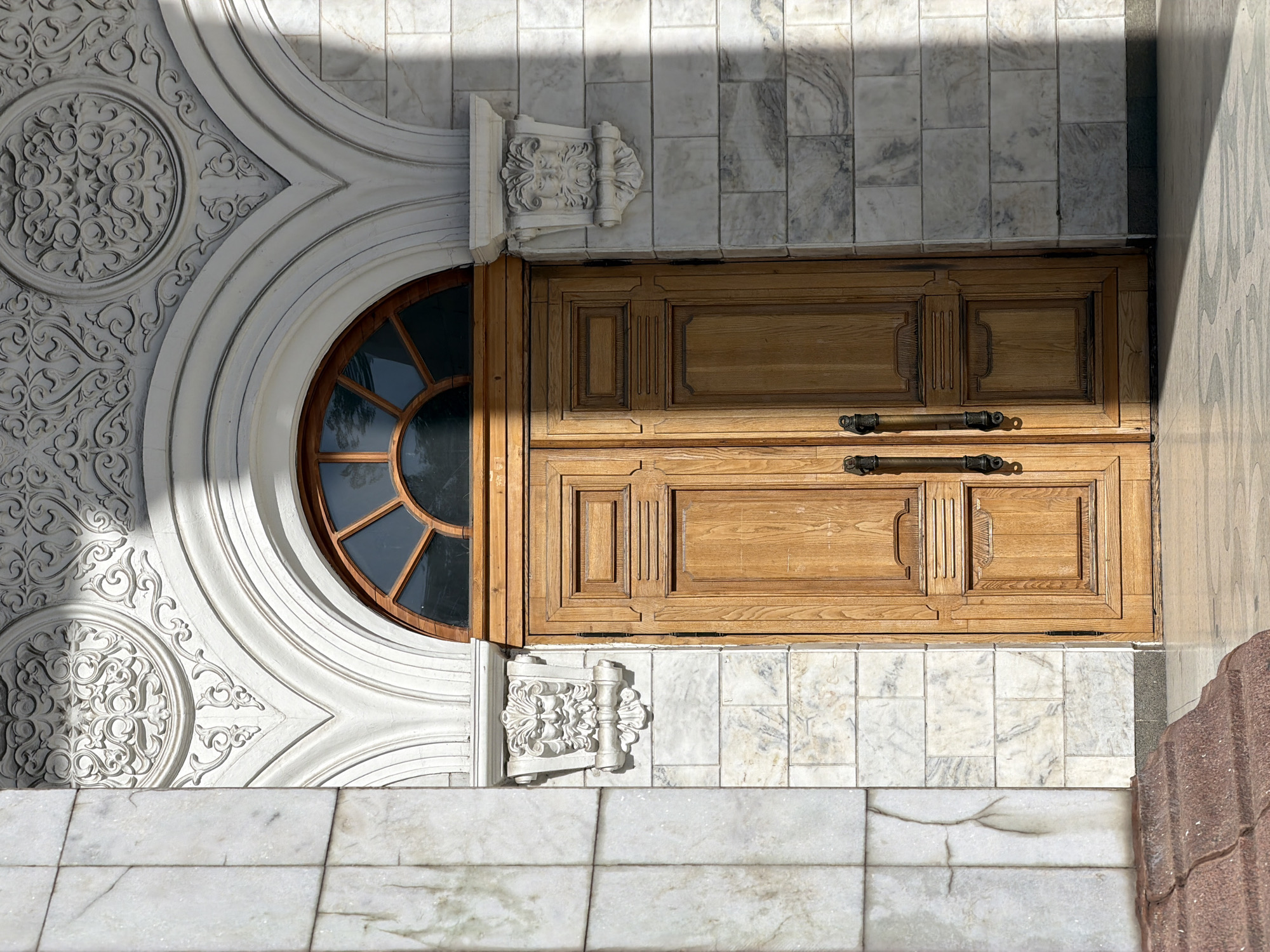 A top-down, daytime shot showcases architectural elements of a building, purportedly located in Bishkek City, Kyrgyzstan, based on the presence of a wooden door, a round window, and ornate marble detailing.
The left side features a white, ornate facade with a circular window. The facade is adorned with floral patterns and curved moldings. The window has a wooden frame and radial spokes, creating a sunburst effect.
Adjacent to the window, a wooden door with two horizontal panels is present. Each panel features decorative raised elements and metal handles.
The backdrop and the ground appear to be clad in white marble tiles with grey veining. Light and shadow play across the surface, adding depth and texture.
A section of red-hued, textured material is present in the lower right corner, seemingly part of a wall or architectural feature.