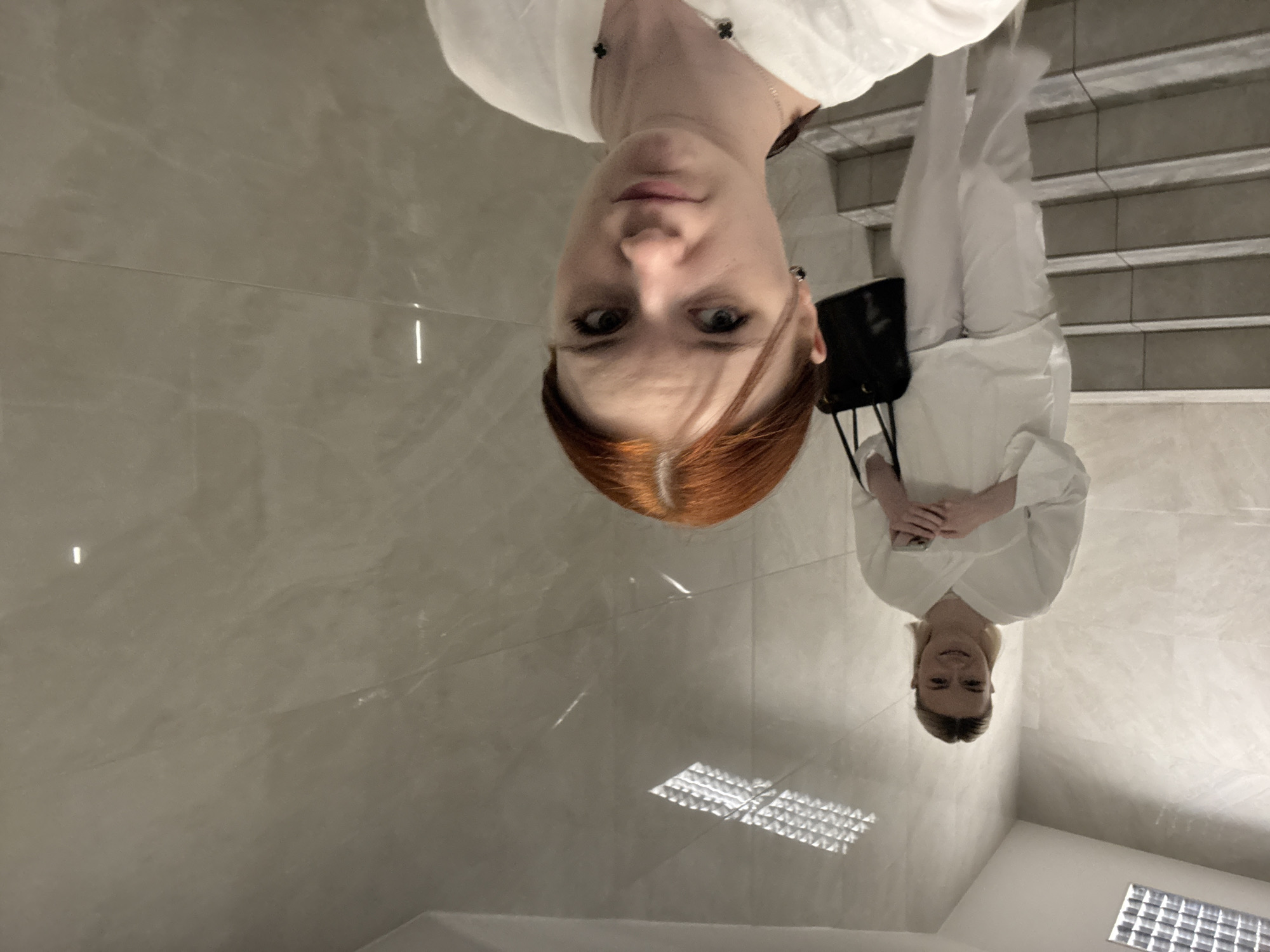 A fair-skinned, red-haired woman is seen upside down in a mirrored reflection. The reflection shows the woman wearing white clothing and standing on stairs with a black shoulder bag. The reflection indicates she is holding a phone. The floor reflects the overhead lights, suggesting a well-lit environment. Based on the overall appearance, this could be a modern interior space. There is not enough information to determine if the image was taken in Saint Petersburg, Russia.