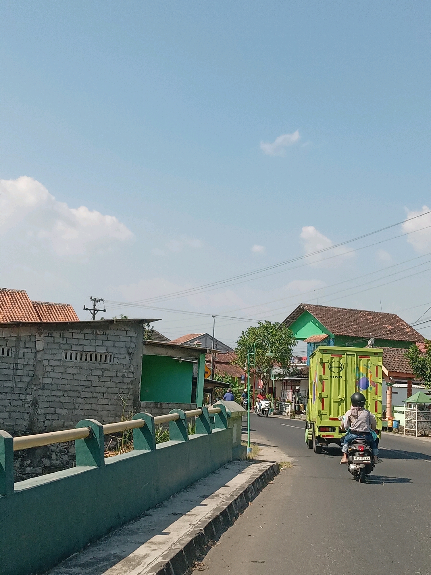 The image depicts a street scene in Srimulyo, Indonesia, on a bright, sunny day with a clear blue sky and scattered white clouds. In the foreground, a dark asphalt road is visible, bordered by a concrete curb and a green, low-rise wall with yellow railings.
On the road, two people are riding a black scooter, traveling away from the camera. The rider, wearing a helmet, has on a light-colored shirt and blue jeans. The passenger behind them is also wearing blue jeans. The scooter has a license plate visible that reads "AB 4655 KK".
Further down the road, a bright lime green truck with a closed cargo box is moving in the same direction. The truck has the brand "HINO" visible on its side, along with colorful circular decals.
On the left side of the road, there is a gray brick building with small rectangular openings in its upper portion and red tiled roofing. Adjacent to this building is a structure painted in a vibrant green.
On the right side of the road, houses with red tiled roofs are visible. One house in the mid-ground is painted green and has a distinctive architectural style. Trees are scattered throughout the scene, providing some greenery. Utility poles and power lines are also present, stretching across the sky. The overall impression is of a typical day in a semi-urban or rural Indonesian setting.