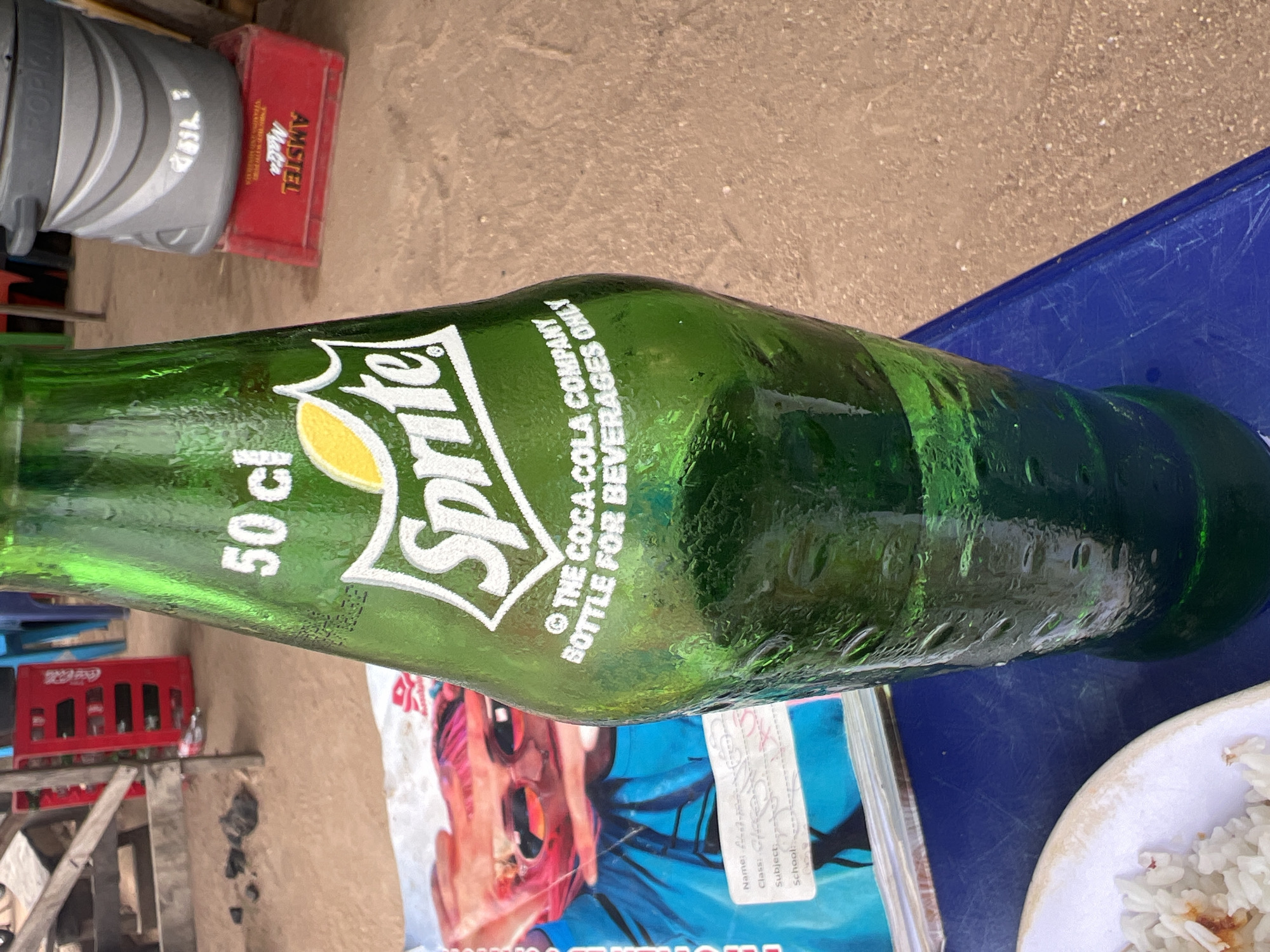 A green glass bottle of Sprite, with the label "50 cl" and "© THE COCA-COLA COMPANY BOTTLE FOR BEVERAGES ONLY", lies horizontally on a blue surface. Adjacent to the bottle is a white plate containing rice. Underneath the bottle is a book with a person on the cover and a partly visible label, which includes the word "School". In the background, there are red crates for Coca-Cola and Amstel Malta, and a stack of metal containers. The scene appears to be outside, likely on a table in Jalingo, Nigeria.
