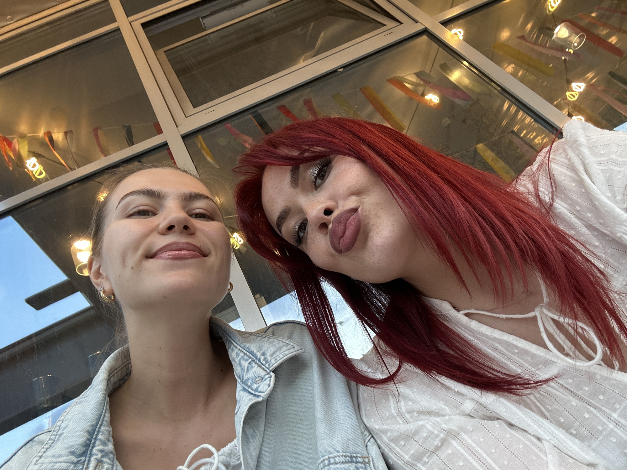 In Tolyatti, Russia, two young women are taking a selfie. The woman on the left has blonde hair pulled back and is wearing a light blue denim jacket. The woman on the right has bright red hair and is wearing a white patterned top. They are positioned in front of a large window with reflections of lights and colorful decorations. The setting appears to be indoors, possibly a cafe or restaurant, with a view of the exterior through the window. The lighting is dim, with warm tones from artificial lights.