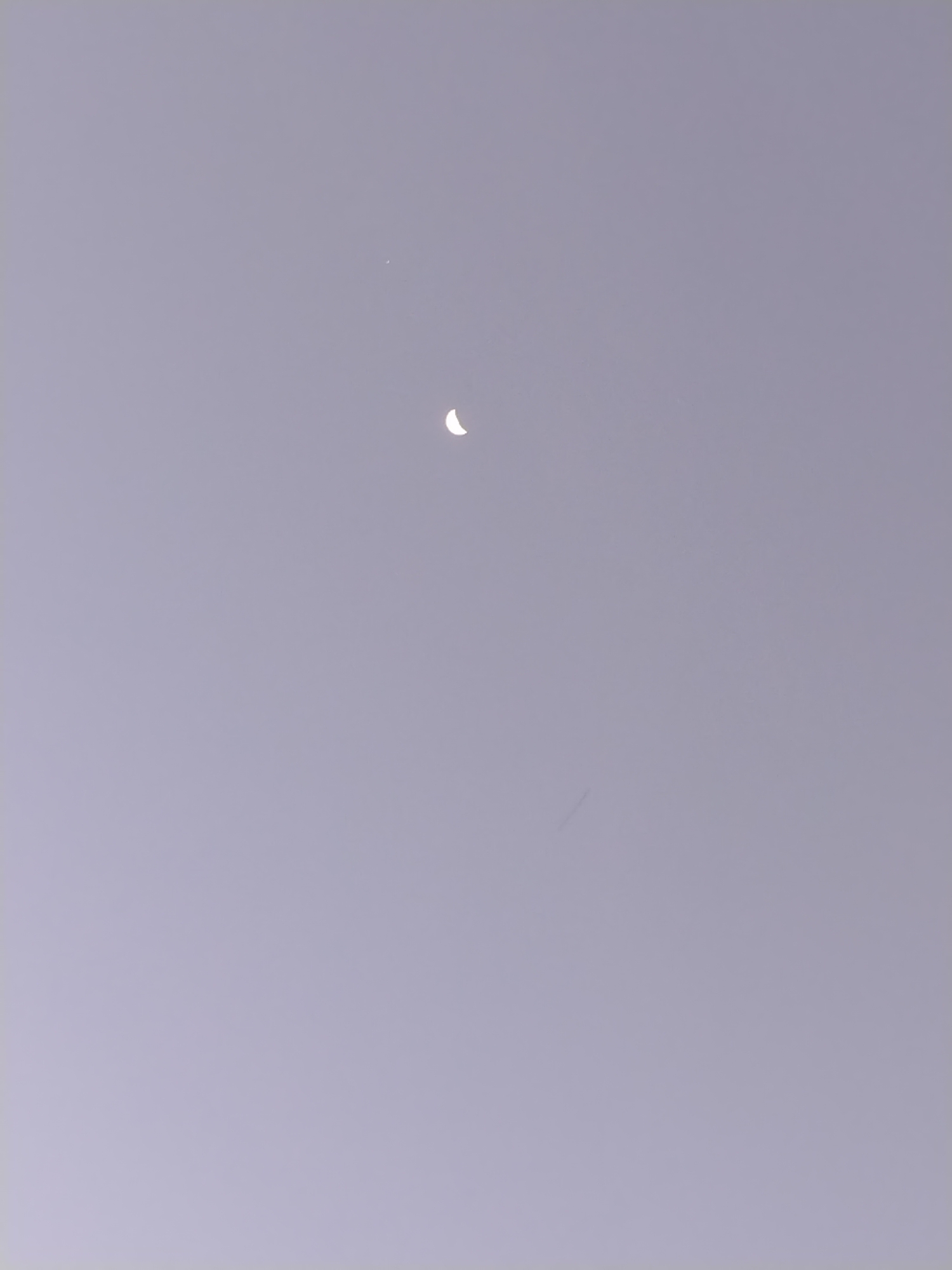The image presents a pale gray sky. A crescent moon is visible in the upper center of the frame, with a small, bright dot (likely a star or planet) positioned slightly above and to the left of the moon. A faint, linear streak can be observed in the lower center of the image, possibly an airplane contrail or an artifact of the image capture. No people, buildings, or other indicators of a city location, like Los Leoncitos, Venezuela, are visible. The scene captures a night sky phenomenon with minimal contextual information.