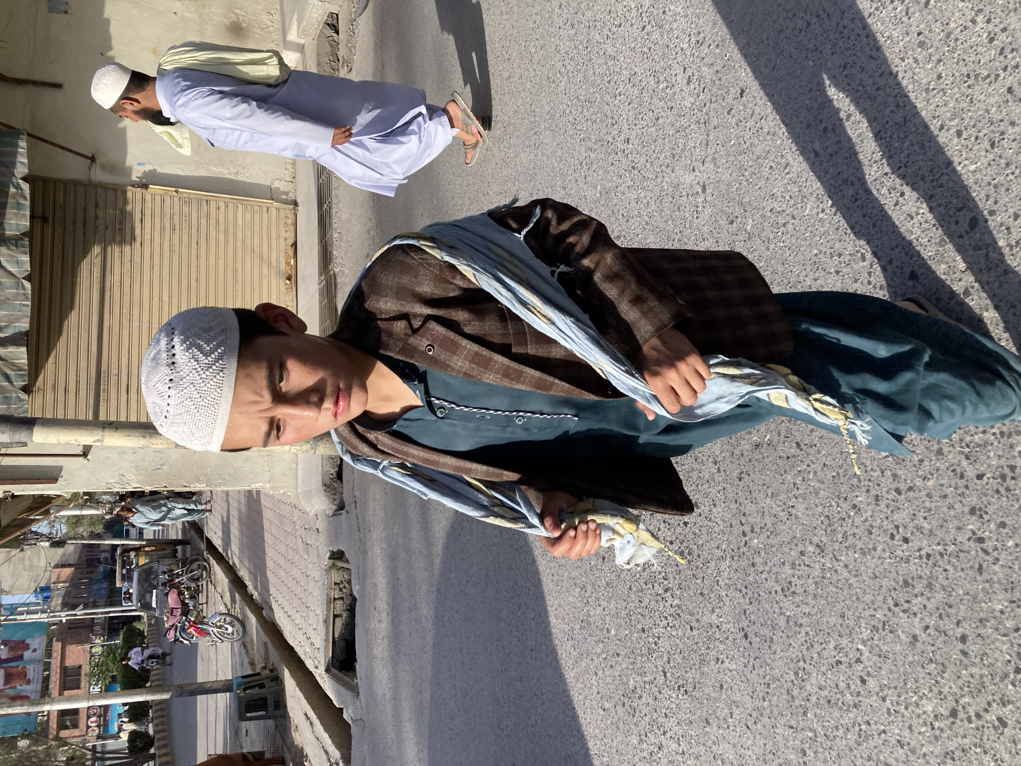 The image depicts an individual in Quetta City Tehsil, Pakistan. The main subject, wearing a white kufi, a dark blue shirt, a brown blazer, and a blue and white scarf, is positioned in the center, partially obstructing the view of a person in the background who is clad in traditional white clothing with a cream-colored shawl. The environment appears to be a street, with buildings visible on the left and shadows cast on the gray road surface. In the background, there are motorcycles and other indistinct vehicles.