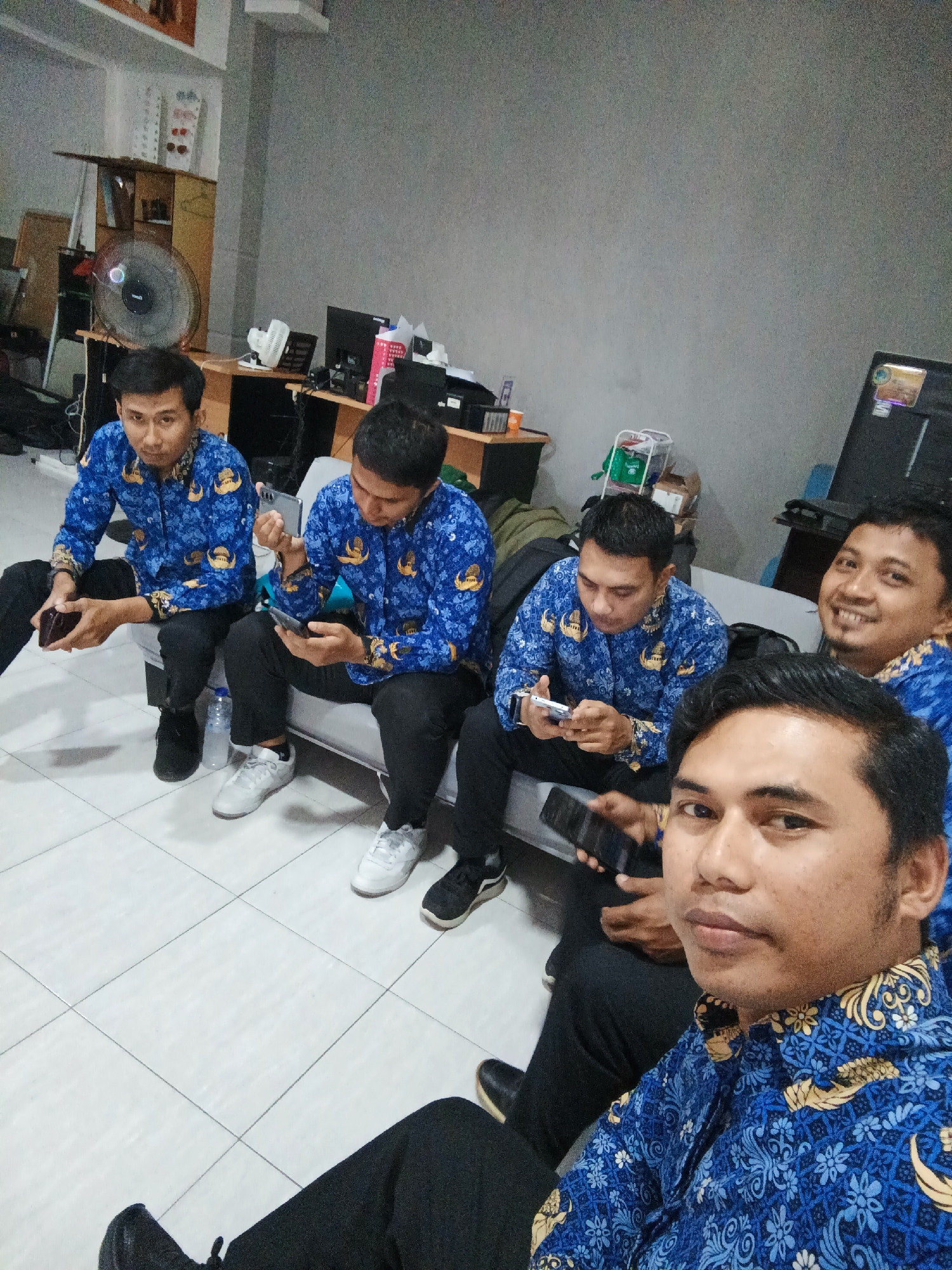 A medium shot captures five adult males in blue Batik shirts with a gold and white pattern, seated on a light gray sofa in what appears to be an office setting. The men are mostly looking at their mobile phones. The individual closest to the camera is in the foreground, facing slightly to the left and looking directly at the camera. He has dark hair and a light complexion. Behind him, to his right, another man is smiling and looking towards the camera. He also has dark hair and a light complexion. To the left of the foreground individual, two men are seated next to each other, both looking down at their phones. The leftmost of these two men is holding a dark wallet in his left hand. The man to his right is holding his phone. Further back, a fifth man is seated on the sofa, also looking at his phone.
The environment includes a light gray wall in the background, with a shelf unit to the left displaying various items including what appears to be a framed picture or diploma, a white fan, and some books. In the middle ground, to the right of the shelf unit, there is a desk with a black computer monitor and other office supplies. A dark gray cabinet or desk is visible in the far background. The floor is tiled with light-colored square tiles. A water bottle is on the floor near the sofa. The overall lighting is moderate, suggesting an indoor setting. The location in the city of Gorontalo, Indonesia, is not discernible from the visual information provided.
