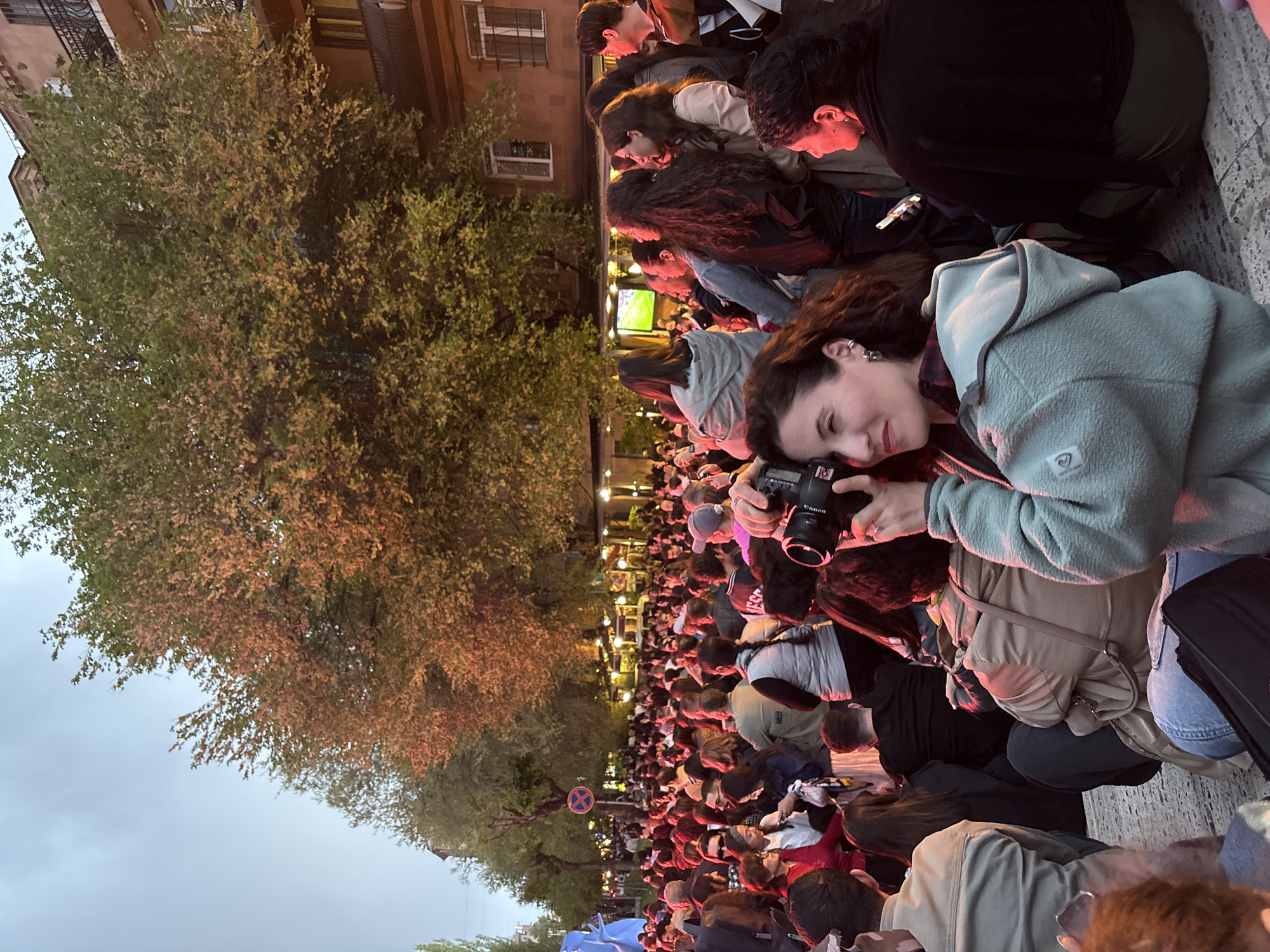 This vertical image depicts a lively evening outdoor gathering in Yerevan, Armenia, with a large crowd of people concentrated on what appears to be a street or public square. The scene is illuminated by artificial lights, suggesting dusk or early evening, with a pale blue-grey sky visible above.
In the foreground, a young woman with dark, curly hair is prominent, holding a black Canon camera with a lens, seemingly capturing the event. She wears a light blue, fleecy hooded jacket over a plaid shirt and has a gentle smile, indicating a pleasant experience. Parts of other individuals, including one in a beige jacket, are visible around her.
Beyond the immediate foreground, a dense crowd extends into the distance, with most individuals facing forward, suggesting they are watching a central attraction. Further along, a large screen is visible, displaying what appears to be a green field, possibly broadcasting a sports event. The crowd and the surrounding trees are bathed in warm light from streetlamps and possibly other event lighting, casting an orange glow on the foliage. The trees, full of green and slightly autumnal leaves, line the left side of the street.
In the background, multi-story urban buildings with windows and balconies can be seen. A circular road sign with a blue background and a red cross, indicating "no stopping," is visible amidst the crowd in the mid-distance. The atmosphere appears to be one of collective engagement and enjoyment during this public viewing event.