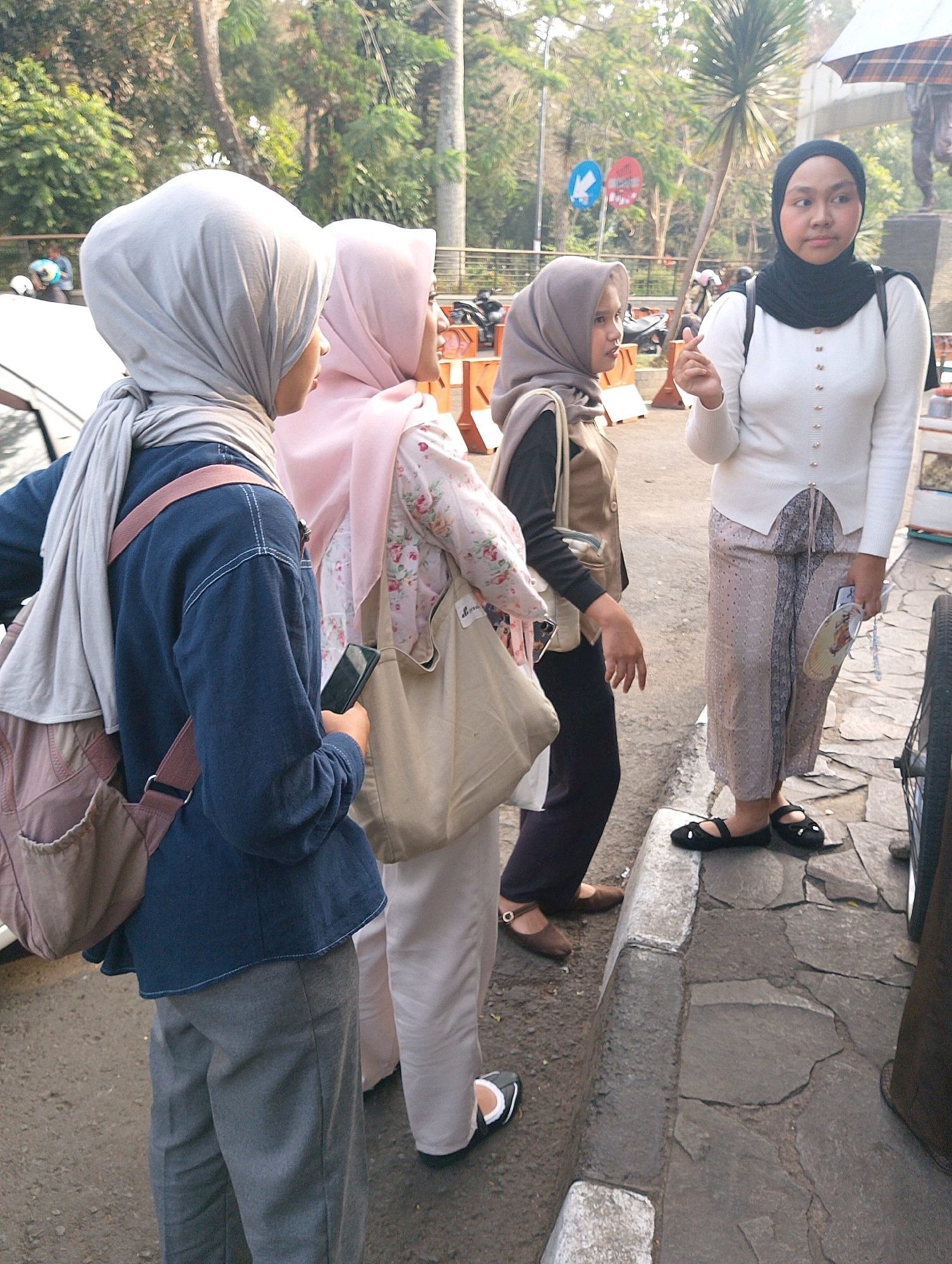 Four young women in hijabs are standing on a cobblestone sidewalk. The woman on the far right wears a white long-sleeved top and a patterned skirt. She holds a folded fan and a phone. The woman next to her, in the center-right, wears a brown vest over a black top and dark pants. The woman to her left wears a light pink patterned top and a light-colored hijab. The woman on the far left wears a dark blue long-sleeved shirt and grey pants, with a light grey hijab and scarf. She carries a pink strap bag and holds a phone. They are standing in front of a stone structure and trees. The background suggests an outdoor, urban setting, possibly a park or a street with pedestrian areas. There are motorcycles and a white vehicle visible in the background. The overall lighting suggests it is daytime. The presence of palm trees and specific vegetation, combined with the architectural style of the stone structure in the background, are consistent with the environment of Bandung City, Indonesia.