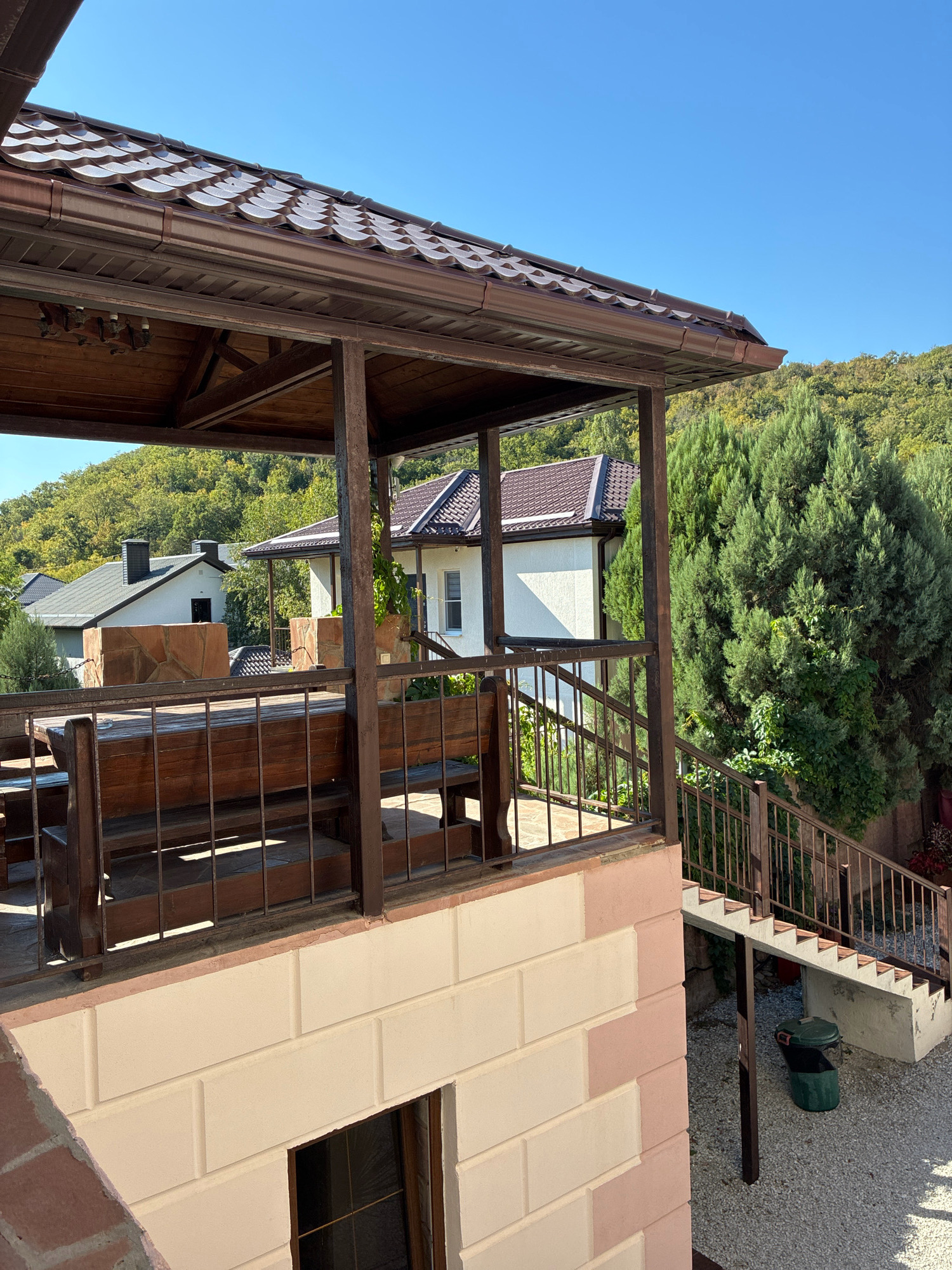 The image depicts an outdoor seating area with wooden tables and benches, overlooking a lush green hillside with several houses. The seating area is covered by a brown tiled roof supported by wooden beams. A metal railing encloses the seating area, and a set of concrete stairs with a metal railing leads down to a gravel path. A green trash can is visible at the bottom of the stairs. The houses in the background have tiled roofs and white walls. The sky is clear and blue. The scene appears to be located in a residential area surrounded by nature, consistent with the landscape of Abrau-Dyurso, Russia. There are no people or visible animals in the image.