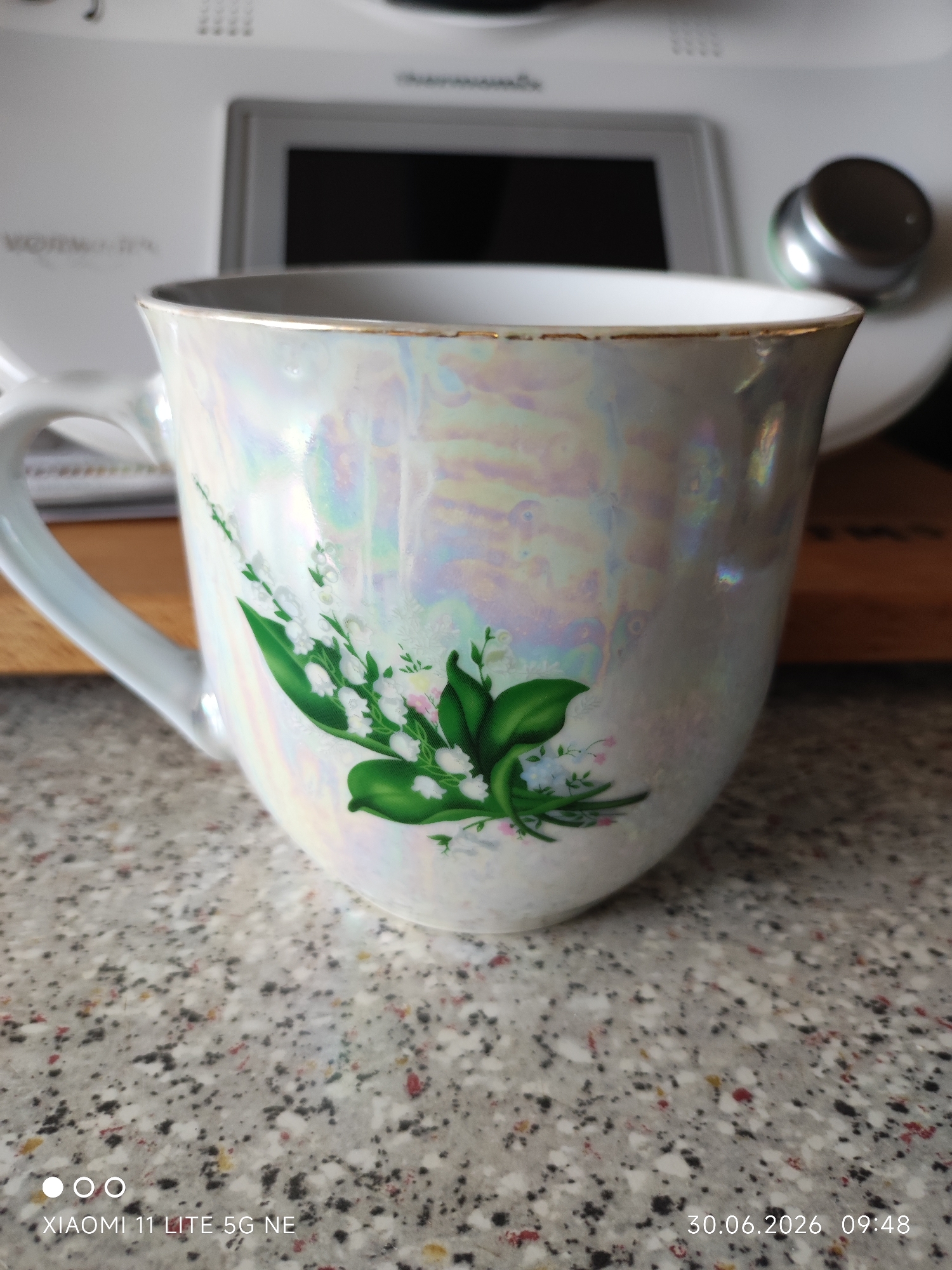 A pearlescent mug with a floral design featuring lily of the valley sits on a speckled countertop. Behind the mug, a portion of a white kitchen appliance is visible, with a black screen and a silver dial. The background suggests an indoor setting. The mug is the primary subject, and the design is the most prominent detail on it.