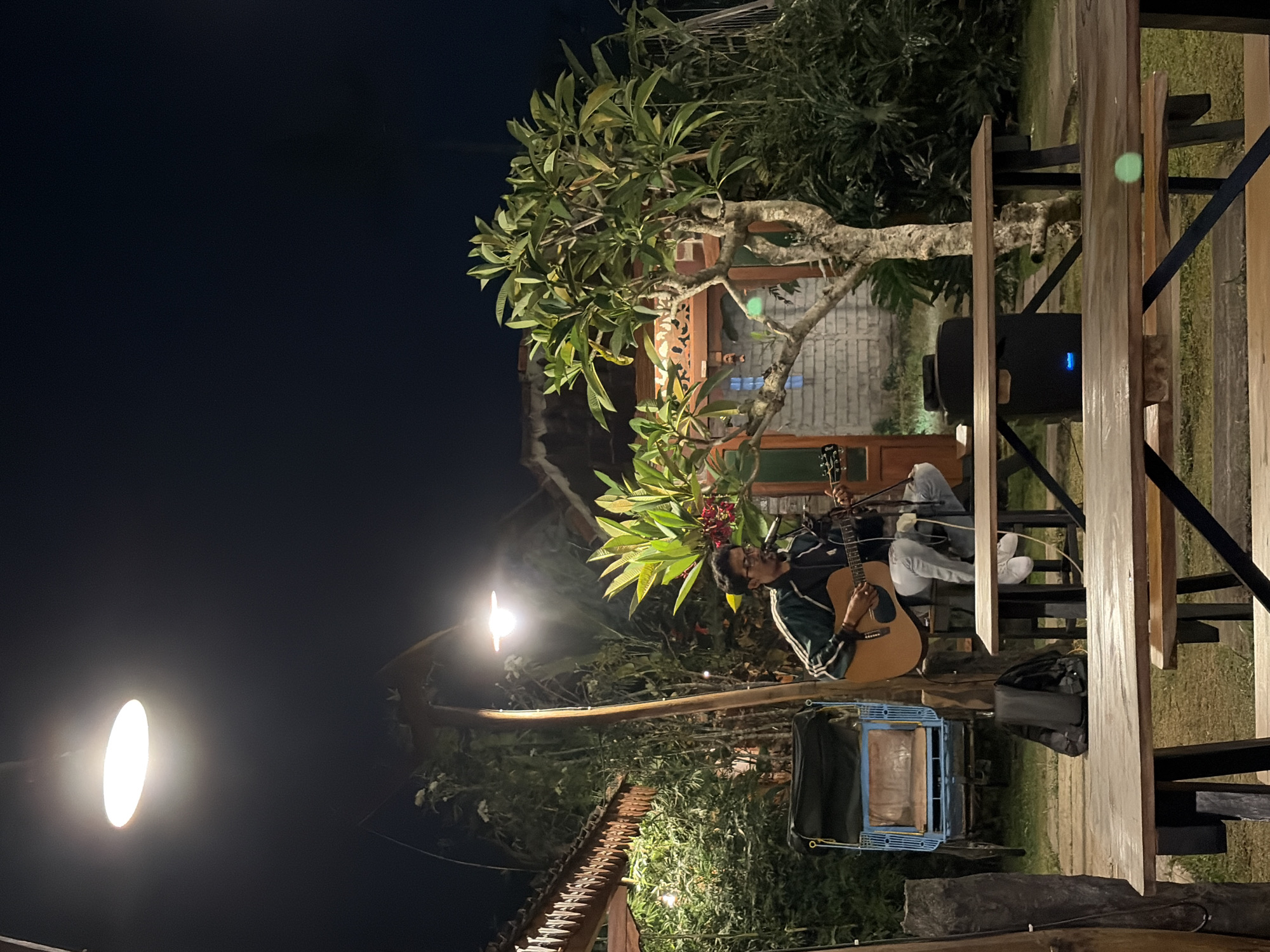 A young man with glasses is playing an acoustic guitar and singing into a microphone. He is sitting on a stool under an outdoor lamp in what appears to be a garden or patio area. The man is wearing a dark green jacket with white stripes on the sleeves, a black shirt, and light-colored pants. A black backpack is visible on the ground behind him, next to a blue and yellow cart. In the background, there is a tree with green leaves and red flowers, a building with a carved wooden facade, and dense foliage. The scene is illuminated by an overhead light and appears to be taking place at night. The location is identified as Campurejo, Indonesia.