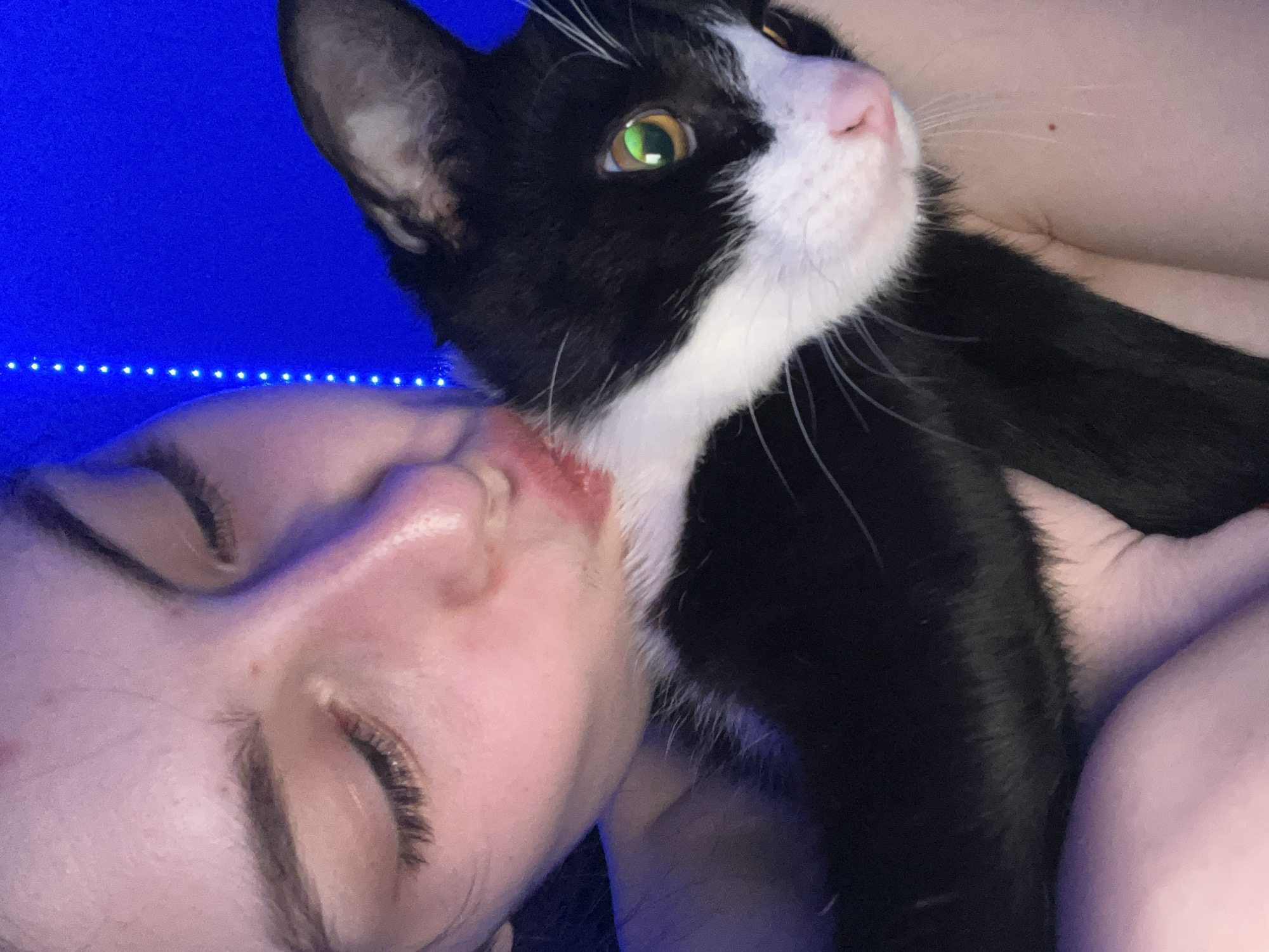 A close-up, high-angle indoor shot captures a young person with their eyes closed, resting their face against a black and white cat. The lighting is dim, with a vibrant blue light source illuminating the background and casting a blue hue on the subjects. The person, whose face is prominent in the bottom left, appears to be sleeping or in a relaxed state. Their eyelashes are visible, and there's a hint of red on their lips or near their mouth, possibly makeup or a mark. The black and white cat, with bright green eyes and pink nose, is looking upwards and to the right, away from the person. Its whiskers are visible, and its fur is a mix of black and white patches. A thin string of blue LED lights runs horizontally across the upper background, suggesting an artificial light source in the room. The overall atmosphere is intimate and peaceful, implying a strong bond between the person and their pet. No visible text is present.