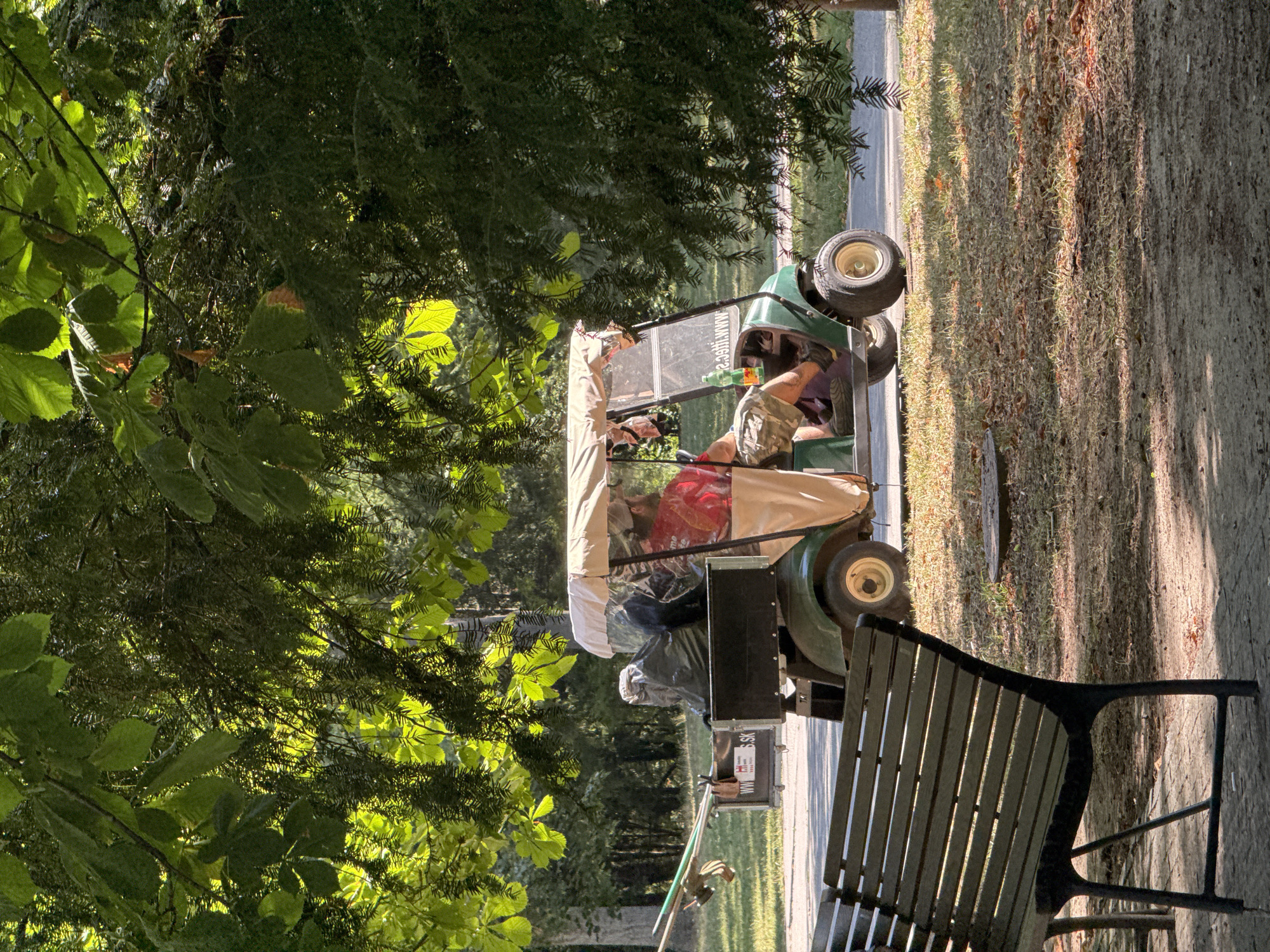 A man in camouflage shorts and a red shirt drives a green golf cart on a paved path, with a trailer attached behind it filled with garbage bags and long tools. The golf cart has a tan canopy and a clear plastic enclosure. In the foreground, a dark wooden park bench is visible. The scene is set in a park with trees and grass, with dappled sunlight filtering through the leaves. The trailer has a sticker on its side that appears to say "WWW.H-SK". The overall impression is of a park maintenance worker operating a utility vehicle.