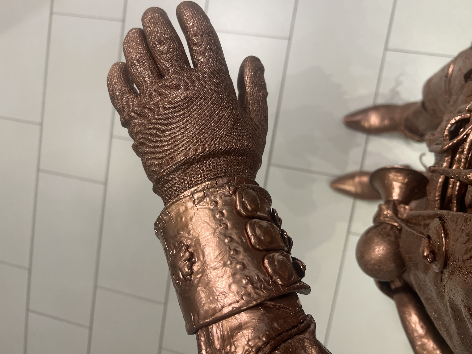 This media file displays a close-up view of a bronze-colored glove and part of an arm. The glove is textured and appears to be made of fabric. The arm is covered in what looks like metallic armor, adorned with rivets and decorative elements. The background consists of a light-colored tiled surface, possibly a wall or floor. The lighting suggests an indoor setting. The image focuses on the detailed craftsmanship of the armor and the texture of the glove. No specific activity or event is depicted, other than the presentation of the object. There is no visible text in the image. The location context of Zakopane, Poland, does not offer any specific visual cues within the image itself.