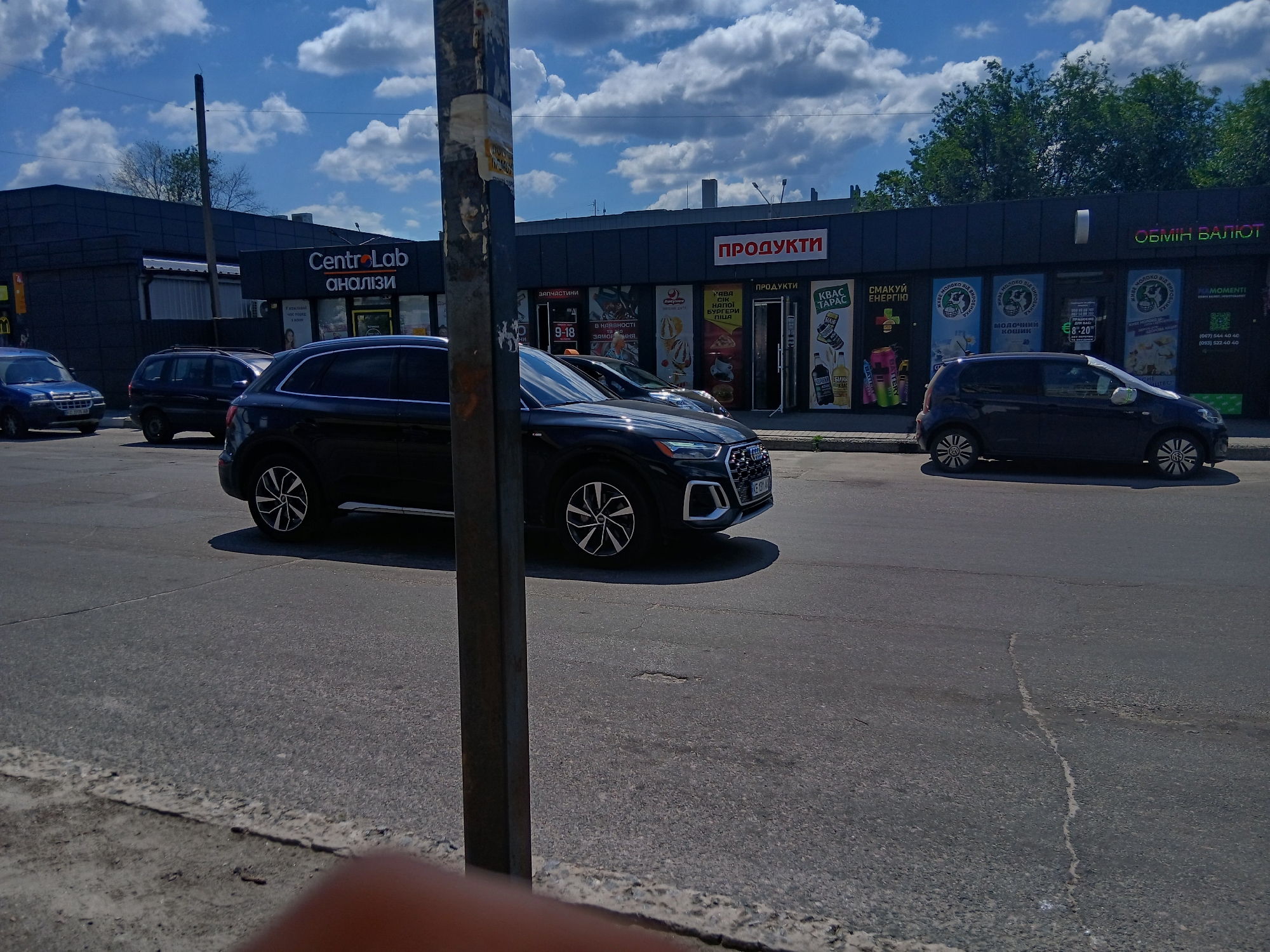 The image depicts a street scene in Dnipro, Ukraine, with a commercial building lining the road. The building features several storefronts with signs indicating various businesses. One prominent sign reads "CentroLab аналізи," suggesting a laboratory or medical analysis center. Another sign displays "ПРОДУКТИ," indicating a grocery or food store. Further along, a sign for "ОБМІН ВАЛЮТ" signals a currency exchange.
Several vehicles are parked or driving on the asphalt road in front of the building. A dark-colored Audi SUV is prominently positioned in the foreground, with its front wheel turned. To its left, a dark-colored SUV and a blue car are visible. A small, dark hatchback is parked to the right of the Audi.
The sky above is blue with scattered white clouds. Trees are visible behind the building, and a metal pole stands near the center of the frame, partially obstructing the view of the storefronts. The ground is paved with asphalt, showing some cracks and wear. The overall impression is that of a typical urban commercial area on a sunny day.