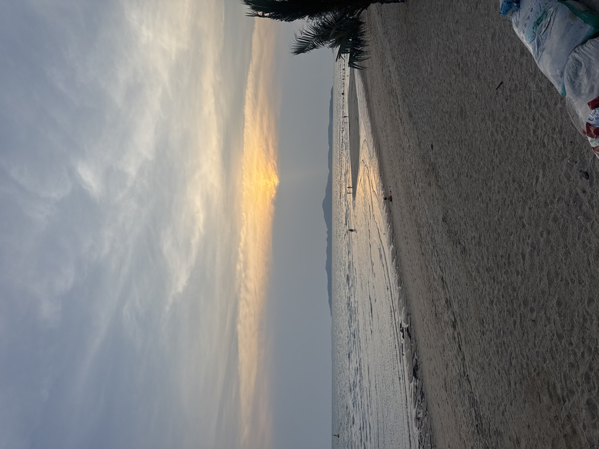 The image captures a serene beach scene at sunset in what is likely Pattaya City, Thailand. The sky, dominating the left portion, features scattered clouds, with a prominent horizontal band of vibrant golden-orange light indicating the sun's position during dusk, contrasting with the softer blue-gray clouds elsewhere.
A wide, sandy beach stretches across the right side of the frame, meeting the calm, reflective waters of the sea. The wet sand near the water's edge shows patterns from receding waves, along with numerous footprints and what appear to be tire tracks. In the distance, low-lying mountains or landmasses are visible across the water.
Several small human figures are scattered along the beach and in the shallow water, casually walking or wading, enjoying the late afternoon ambiance. A distinct individual is seen walking along the distant waterline. A small, bright yellow buoy or marker floats in the middle ground of the water.
On the far right side of the beach, a pile of large sacks is visible, some white or light blue, one with red and white stripes. Some faint, illegible text can be seen on the white sacks. A dark silhouette of a palm tree frond partially frames the upper right corner of the scene. The overall mood is peaceful and relaxing, depicting a tranquil evening on a tropical shore.
