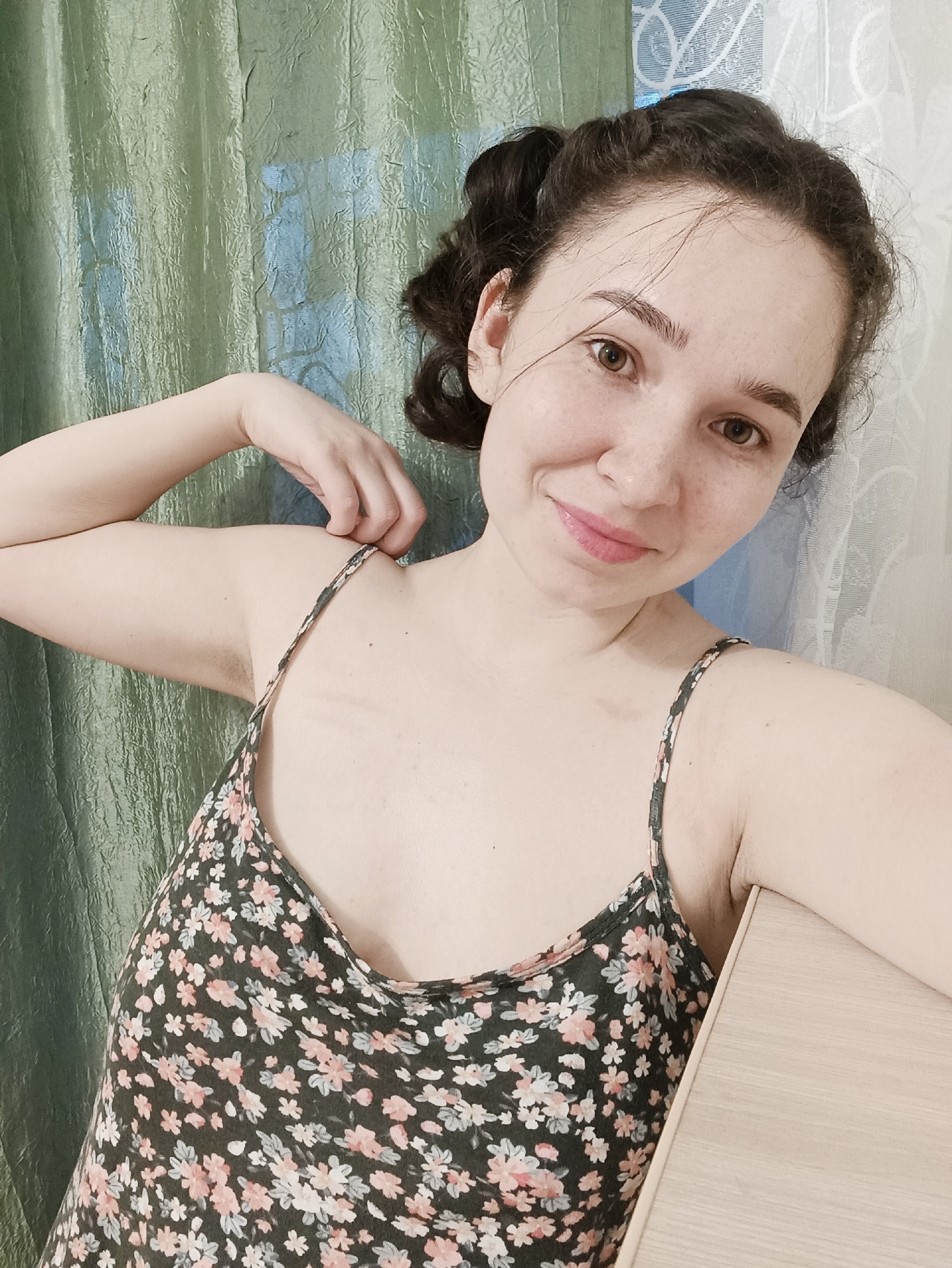 A young woman with brown hair styled in an updo and freckles is posing for a selfie indoors. She is wearing a floral print spaghetti strap top. Her left hand is holding the strap of her top, while her right arm rests on a light-colored surface. The background consists of a textured green curtain on the left and a white wall on the right. There is no indication of the city of Izhevsk or any specific location.