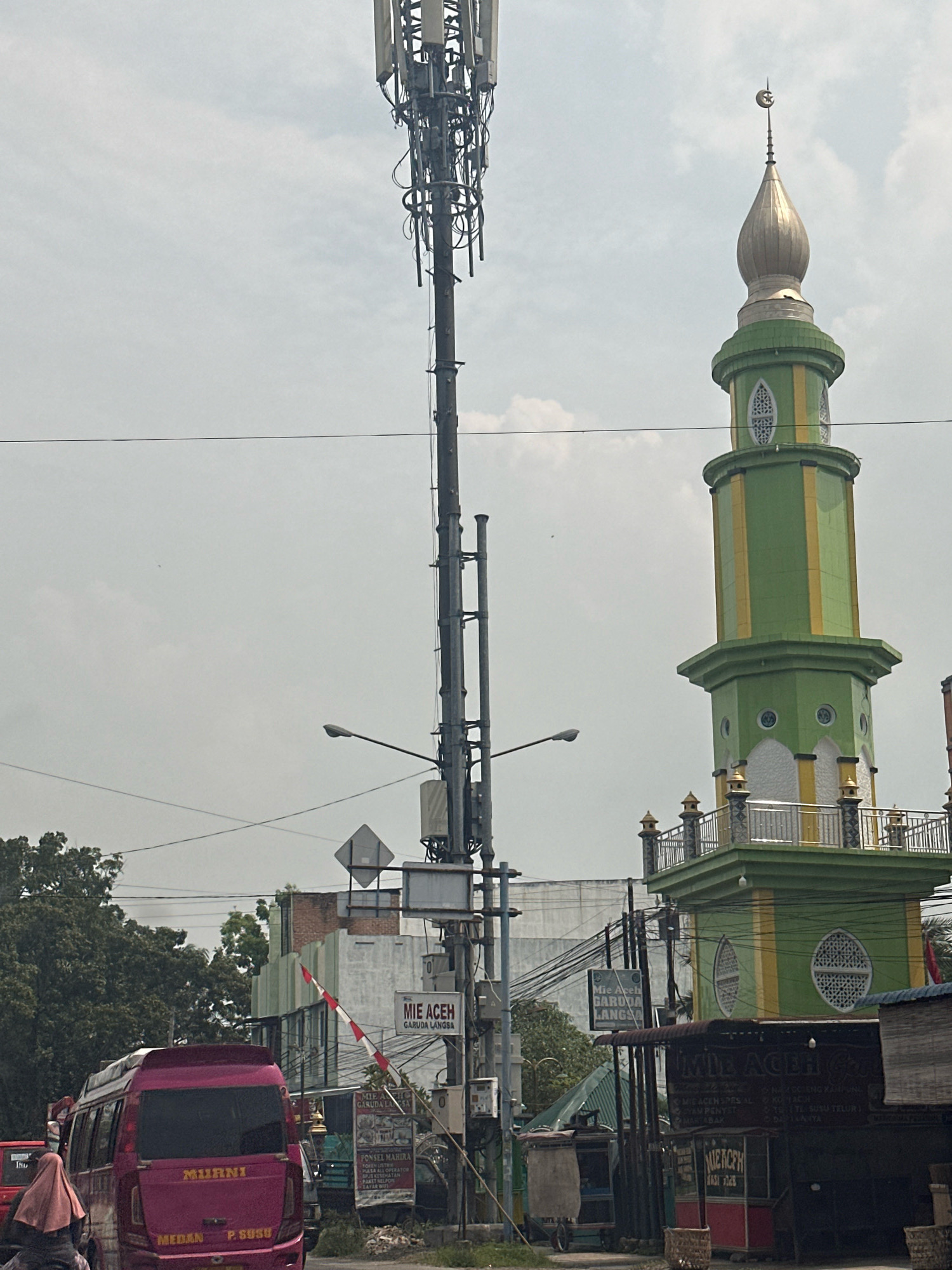 The image depicts a roadside scene in the city of Binjai, Indonesia. Dominating the left side of the frame is a tall telecommunications tower with numerous antennas and cables. To the right, a green and yellow minaret of a mosque rises against a cloudy sky, topped with a crescent moon and star. In the foreground, a pink minivan with the text "MUARNI MEDAN P. SUSU" is visible. A person wearing a pink hijab is partially visible on the left. The background features buildings, trees, and streetlights. A red and white barrier gate is partially visible, suggesting a controlled access point. Several signs in Indonesian, including "MIE ACEH GARUDA LANGSA," are present, indicating local businesses. Power lines crisscross the sky. The overall impression is of a busy urban environment with a mix of modern infrastructure and cultural landmarks.