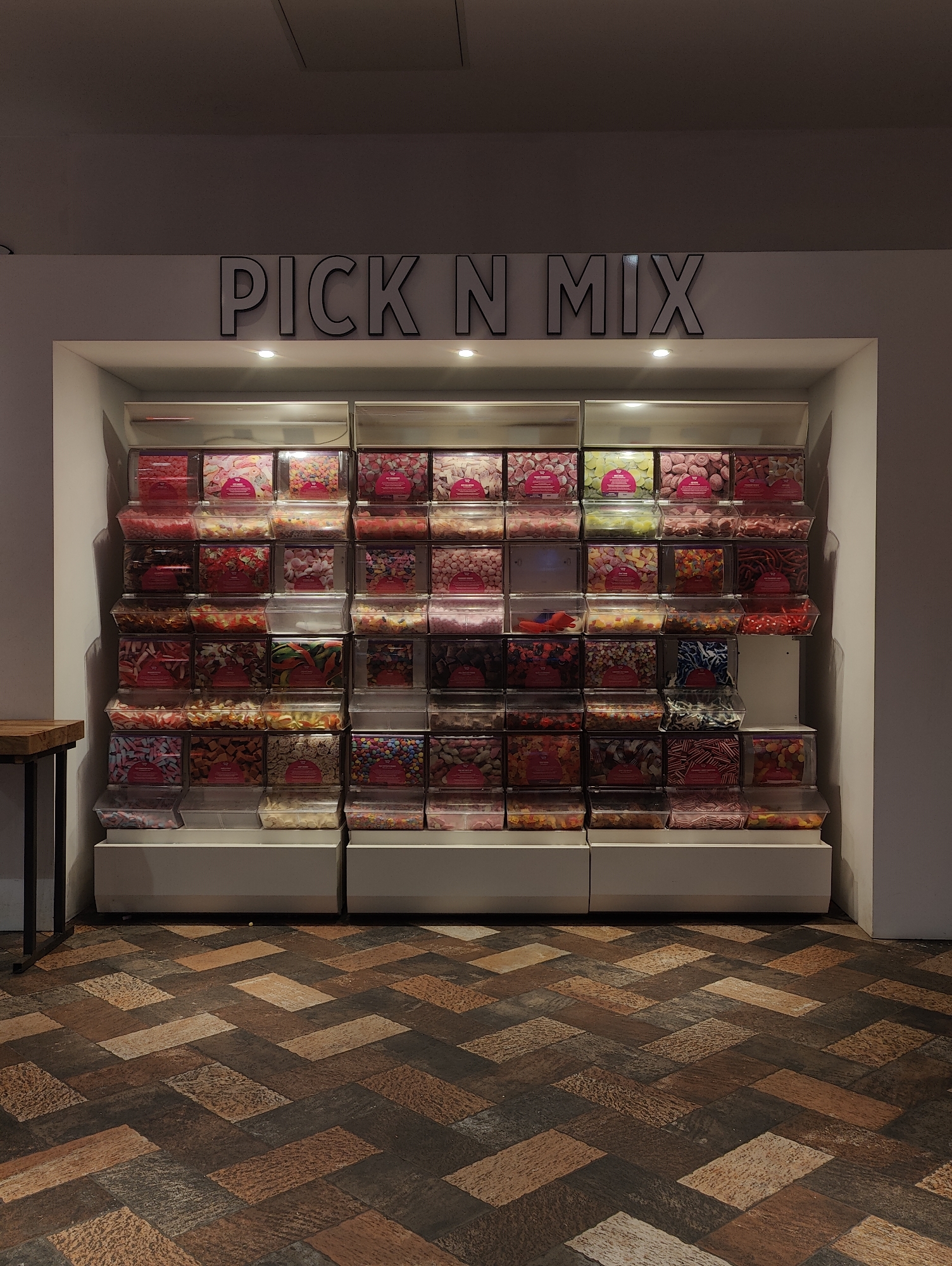 The image depicts a "Pick N Mix" candy display, likely in a retail setting, featuring numerous clear plastic containers filled with various types of candies. The display is set against a white wall, with the words "PICK N MIX" in dark, outlined letters above the candy containers. Small spotlights illuminate the sign. The display itself is built into a recessed area of the wall, with white shelves holding the candy containers. The floor is covered with a herringbone pattern of brown and tan tiles. A portion of a wooden table with black legs is visible on the left side of the frame. Given the presence of the "Pick N Mix" display and general retail aesthetic, the location is likely a candy shop or section within a larger store in the city of Ipswich, United Kingdom.