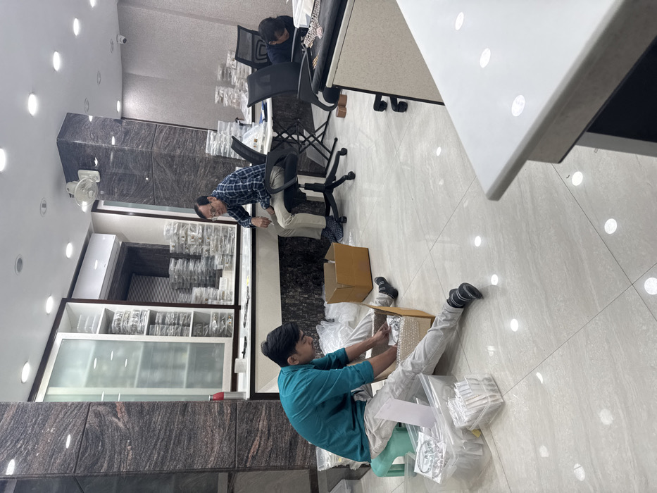 The image shows an indoor commercial workspace, located in Kolkata, India. The area features light-colored, polished tile flooring and columns clad in dark, mottled granite-like material. The walls are predominantly white, equipped with recessed ceiling lights and a white wall-mounted fan. An air conditioning unit is visible on the upper wall.
Three individuals are present. In the foreground-left, a young male, dressed in a teal long-sleeved shirt and light-colored pants, sits directly on the floor. He is actively engaged in handling small, individually packaged items within an open brown cardboard box. A green plastic stool and a clear plastic bin containing more packaged goods are beside him.
In the mid-ground-right, an older male, wearing a blue and white plaid long-sleeved shirt, light-colored pants, and glasses, sits on a black office chair. He is looking towards the left, appearing to observe the activities in the foreground.
In the background-right, a third individual, whose back and head are partially visible, is leaning over a desk, seemingly engaged in work with items on its surface.
The workspace contains white wall cabinets, some with frosted glass doors, and open shelving heavily stocked with numerous small items packaged in clear plastic wrapping. Several black office chairs and white desks with black bases are also present. A red water bottle is visible on a counter in the background.