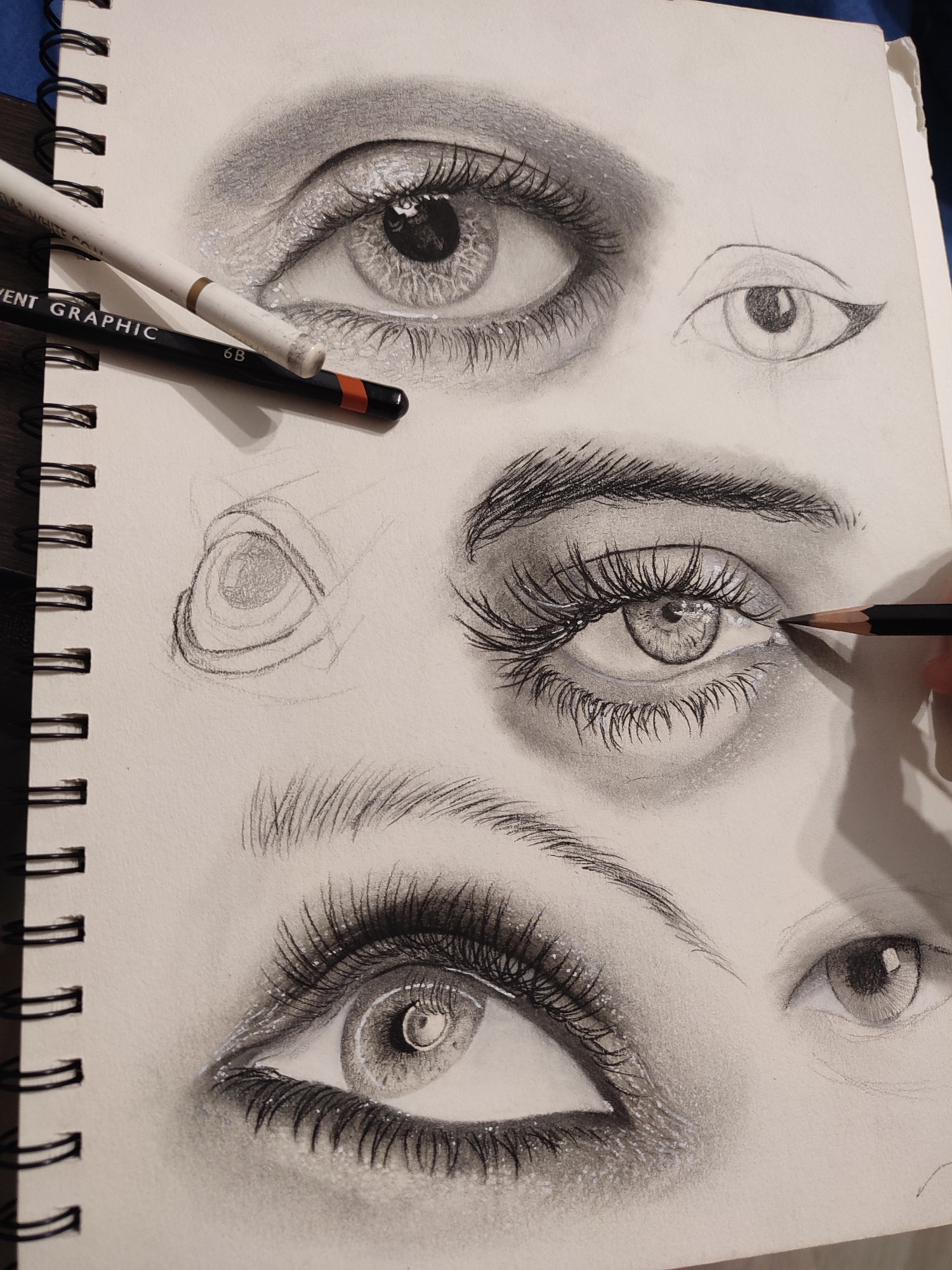 A drawing of several eyes on a sketchpad is shown. The sketchpad is spiral-bound with black rings. Two pencils are resting on the sketchpad; one is white and labeled "CLEAR WHITE COL", and the other is black with an orange tip and labeled "6B". The drawing itself is in pencil and features various stages of completion for several eyes, with one eye being drawn in detail with prominent eyelashes and shading. Another eye is lightly sketched, and there are also several preliminary sketches of eyes.