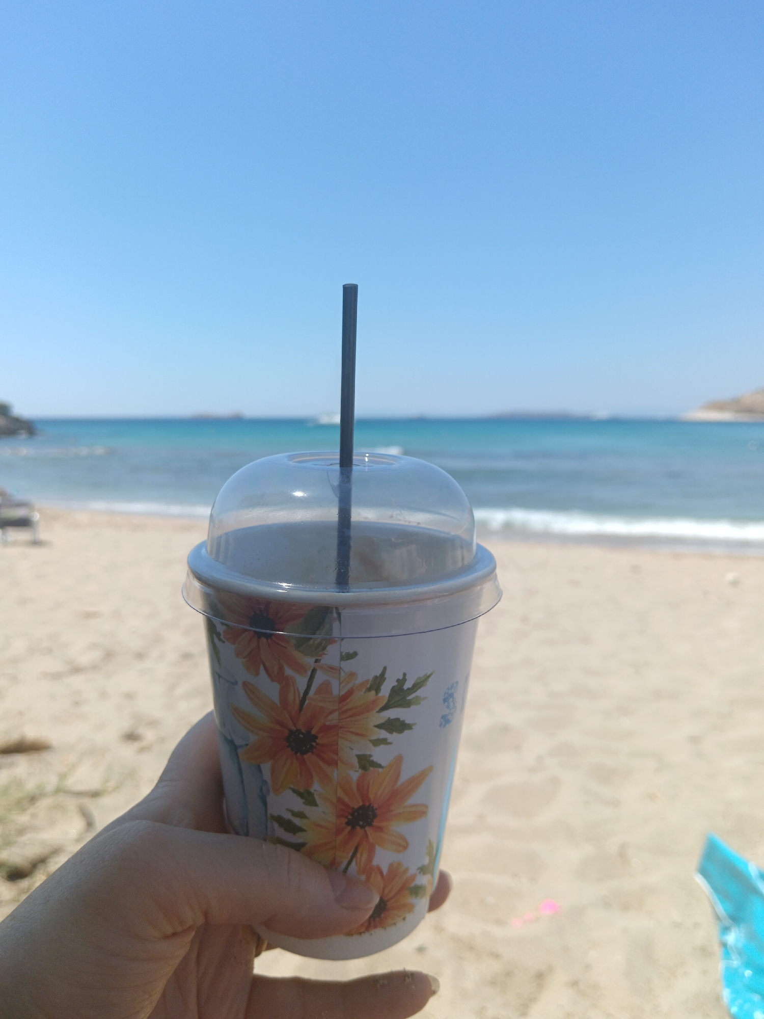 In Agia Marina, Greece, a hand holds a paper cup with a floral design and a clear plastic lid, from which a black straw protrudes. The cup contains a light-colored beverage. In the background, a sandy beach transitions to the turquoise waters of the sea, with gentle waves breaking on the shore. The sky above is a clear, pale blue. In the distance, a hazy landmass is visible on the horizon. A blue beach toy is partially visible on the sand in the lower right corner.