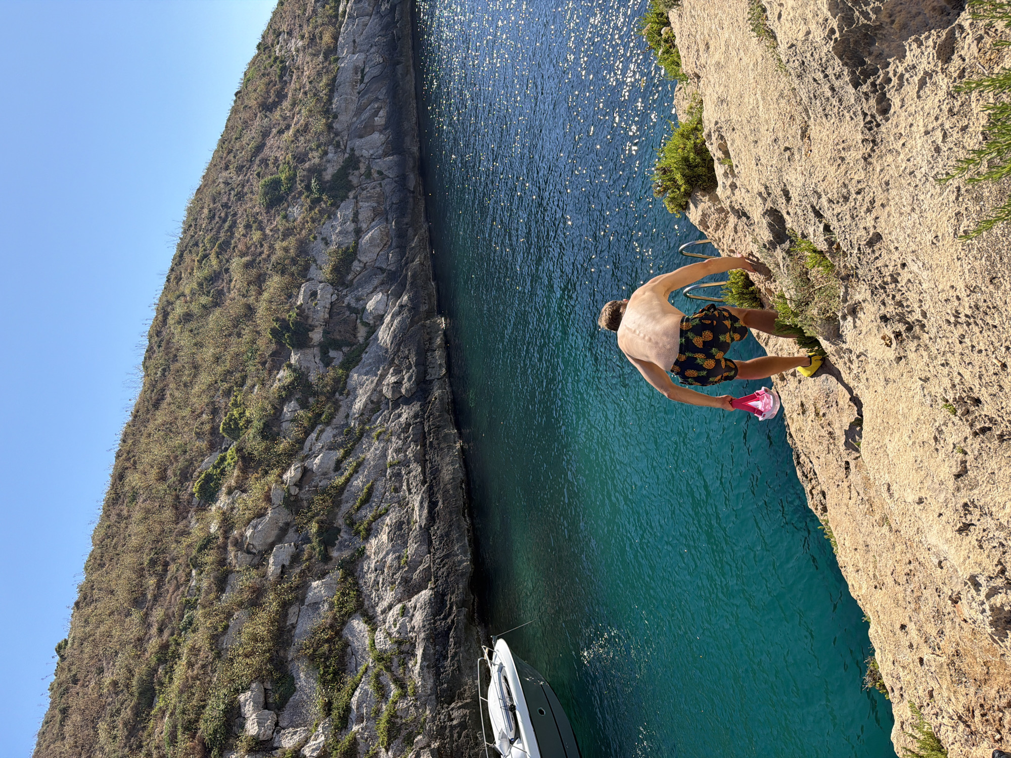 In Xlendi, Malta, a shirtless man wearing pineapple-patterned swim shorts and yellow shoes climbs down a rocky incline towards a ladder with chrome rungs, holding a pink snorkel mask and goggles. He is positioned on a tan, rugged cliff face overlooking a deep, clear turquoise body of water that sparkles under a bright, clear blue sky. To the left, a white motorboat is moored in the water, its bow facing the shore. The rocky cliffs rise steeply on the opposite side of the bay, covered in sparse, dry vegetation. The scene depicts a person preparing to enter the water for swimming or snorkeling.