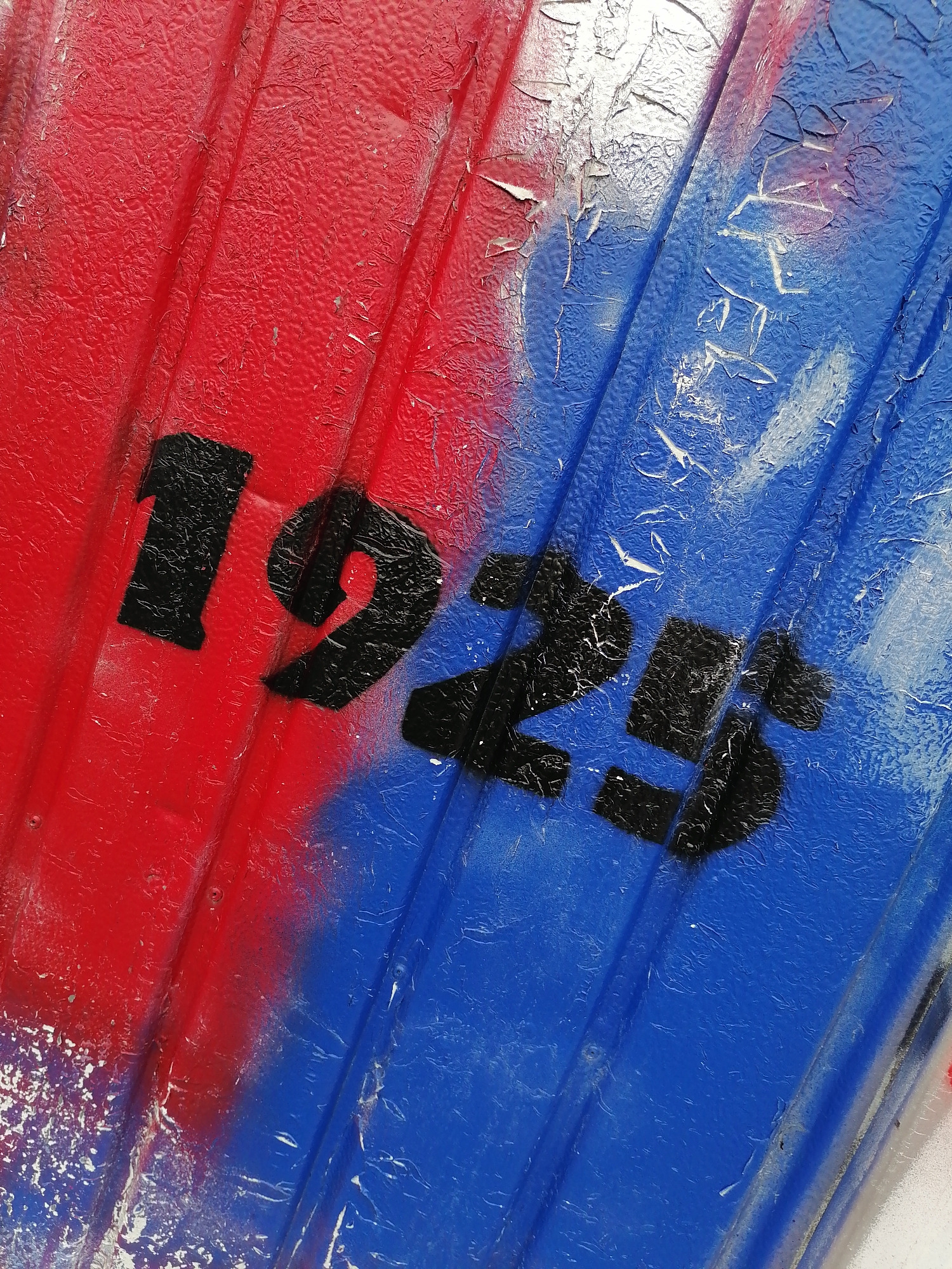 The image is a close-up, diagonal shot of a corrugated surface painted in alternating sections of red and blue. The number "1925" is spray-painted in black over the red and blue sections. The paint is peeling and cracked, revealing a metallic surface underneath, suggesting age and weathering. This object is located in Unterhaching, Germany. No people or actions are visible in the image.