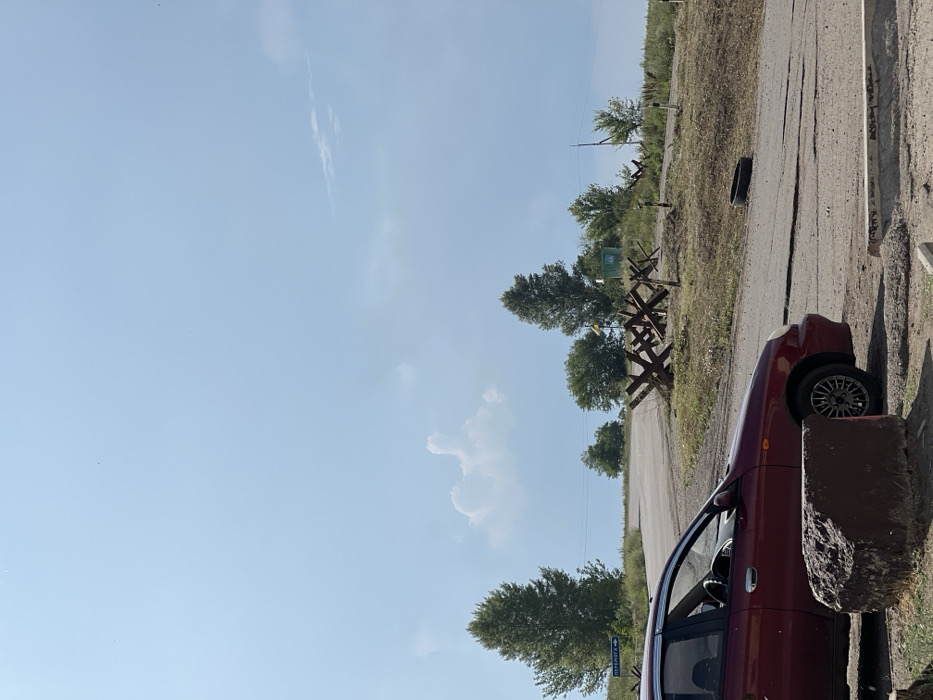 The image depicts a roadside scene in Shestakove, Ukraine, under a clear, bright blue sky with scattered white cumulus clouds, one of which exhibits a distinct, elongated vertical formation. In the lower-left foreground, the front and mid-section of a stationary maroon four-door sedan are visible, parked on an asphalt road with visible cracking. The vehicle faces towards the right. A large, dark, rough-textured block of rock or concrete rests in the immediate foreground, partially obscuring the front of the sedan.
To the right of the road, on a dry, grassy slope, a line of approximately six dark-colored metal anti-tank obstacles, commonly known as Czech hedgehogs, are arranged in a defensive pattern. Further right on the slope, a single black vehicle tire is discarded on the ground. A blue road sign with white Cyrillic text reading "ПЕРЕМОГА" (Peremoha) and a right-pointing arrow is situated on the left side of the road, behind the sedan. The road appears to ascend and curve to the right in the mid-ground. Deciduous trees with green foliage are distributed across the scene, particularly on both sides of the road and in the background. No individuals are present. The overall environment suggests a rural or semi-rural setting under daylight conditions.