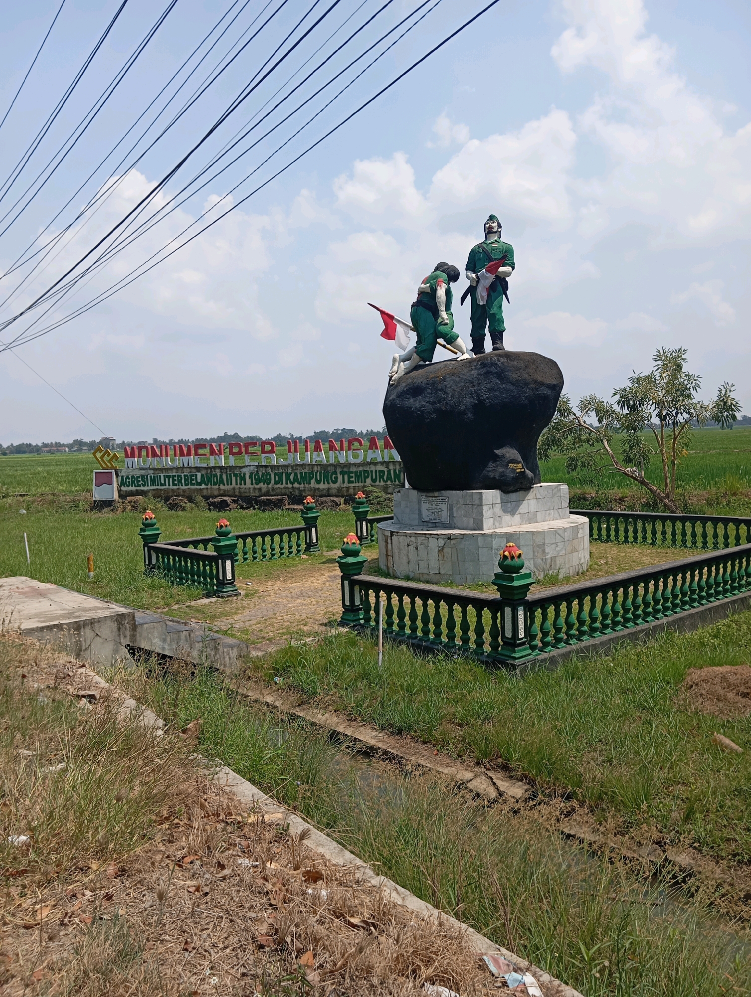 The image shows a monument in Leman Benawi, Indonesia, dedicated to "AGRESI MILITER BELANDA II TH 1949 DI KAMPUNG TEMPURAN." The monument features two statues of soldiers in green uniforms, one standing and the other kneeling, positioned atop a large dark rock. The standing soldier holds a flag, and the kneeling soldier appears to be holding a rifle. The statues are part of a larger monument with a white marble base. A green metal fence surrounds the monument, adorned with decorative orange elements on its posts. In the background, there is a large banner with red lettering that reads "MONUMEN PERJUANGAN." The surrounding environment consists of a grassy field, a small canal, and a bright blue sky with scattered clouds. Telephone wires are visible in the upper portion of the image.