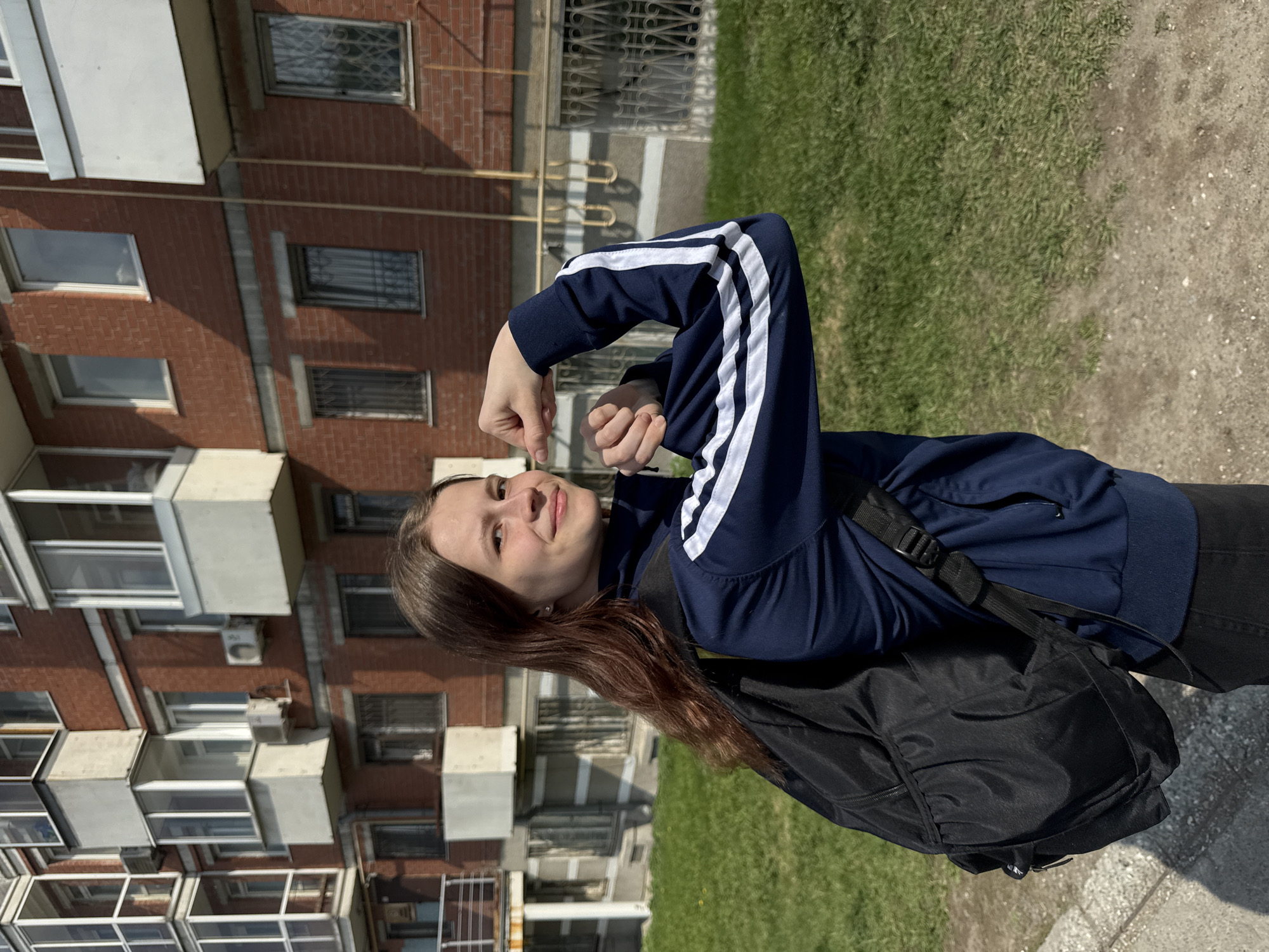 A young woman stands outdoors on a sunny day, posing for a photo. She wears a dark blue jacket with white stripes on the sleeves and a black backpack. Her right hand is near her face, making a gesture. The backdrop includes a residential building constructed of red brick and white concrete. There is a patch of green grass to the right. The setting is likely in an urban area, possibly in Perm, Russia, based on the architecture of the building.