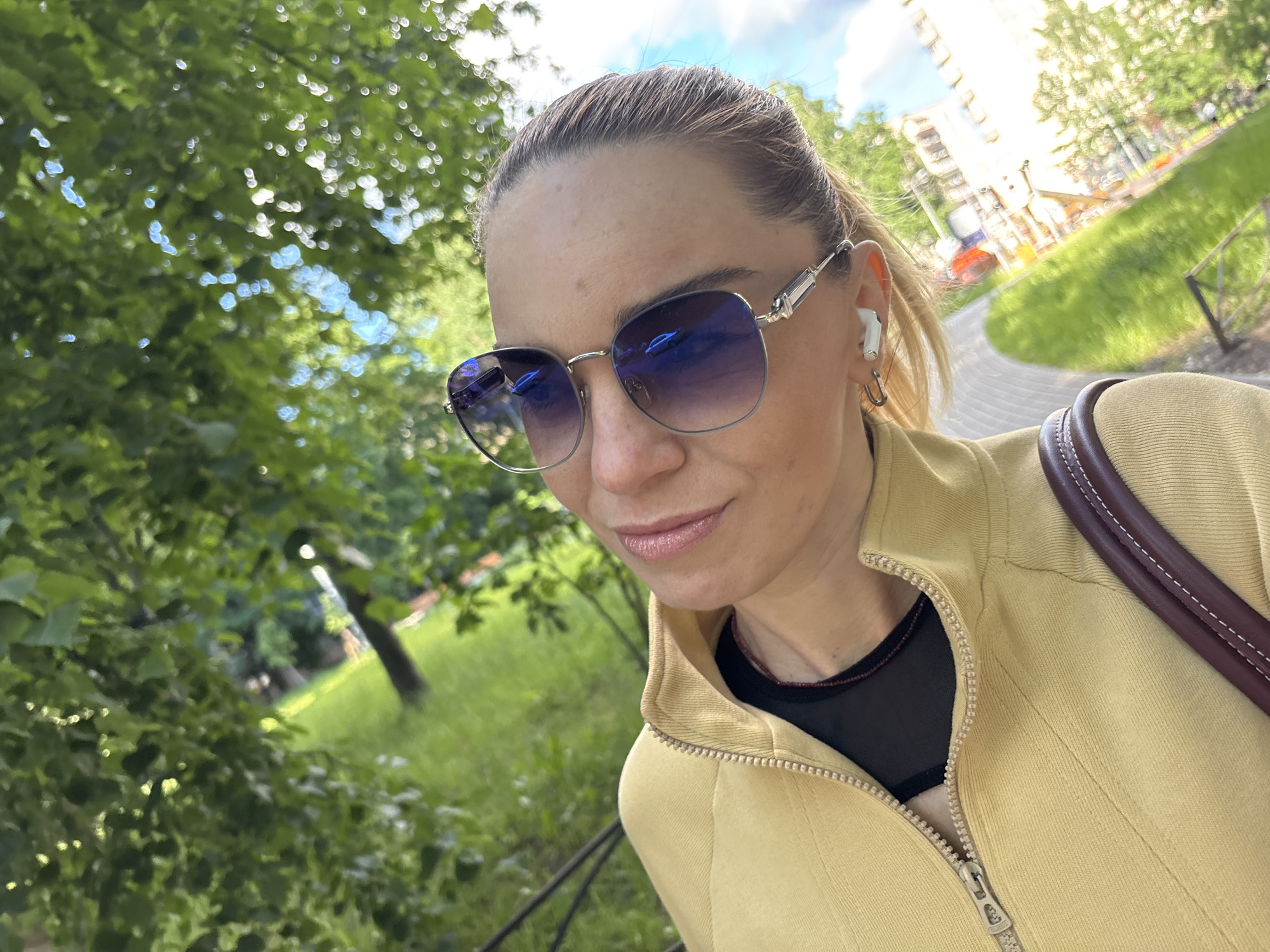 A woman, seen from the chest up, poses for a selfie outdoors. She is wearing sunglasses, a yellow jacket zipped halfway up over a black top, and white earbuds. Her hair is pulled back. The background is a mix of green foliage, grass, and a building in the distance, suggesting an urban park or street.