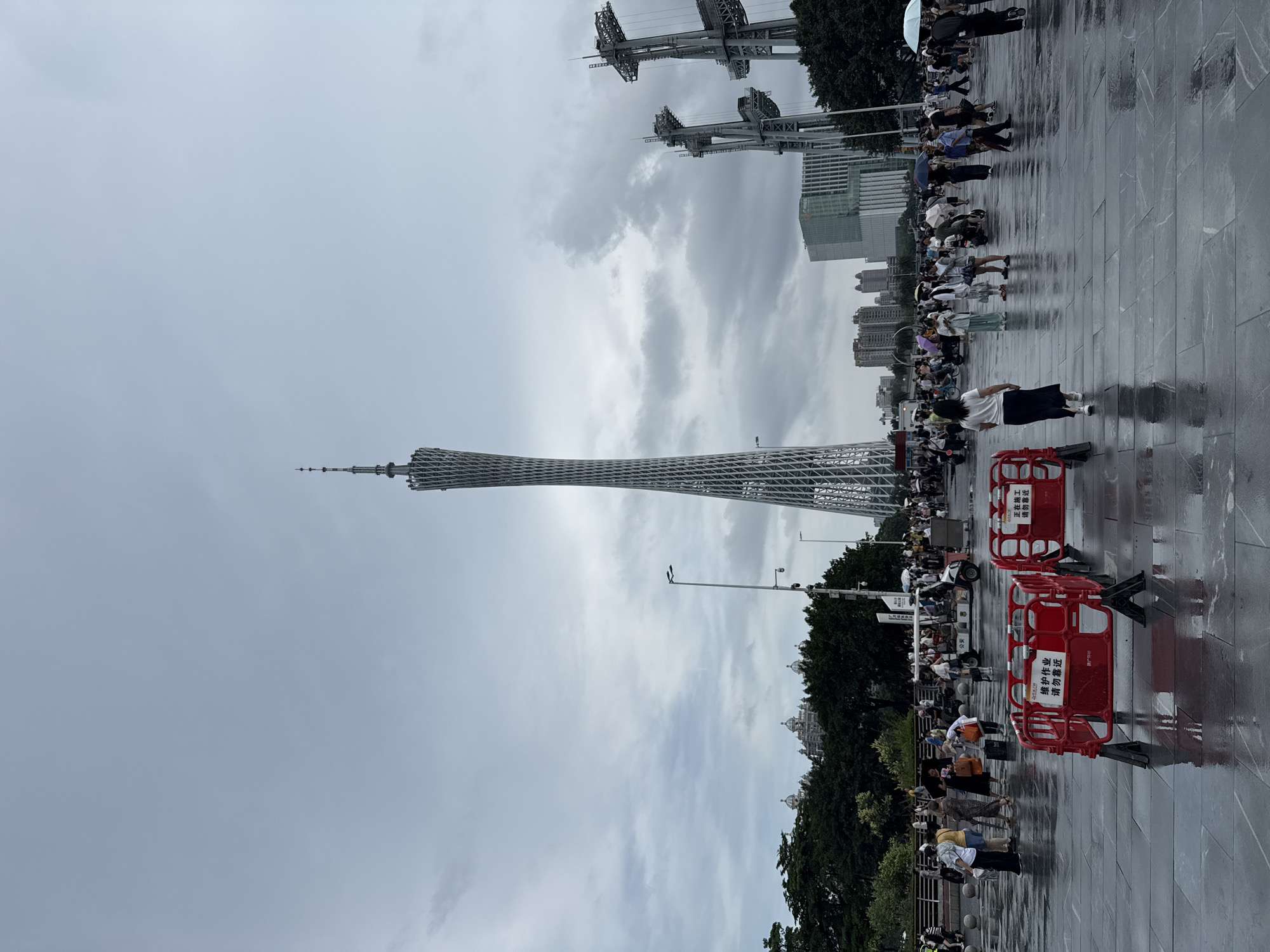 The image shows a wide-angle view of the Canton Tower, a prominent landmark in Guangzhou, China, located in the Tianhe District. The sky is overcast with heavy clouds. In the foreground, a paved plaza area is wet and reflective, indicating recent rain. Two red temporary barriers with white signage are placed on the plaza. The barriers have Chinese text that translates to "Continuing work, please stay away." Many people are walking around the plaza, some carrying umbrellas, suggesting it might have rained or is about to rain. Some are dressed casually, while others appear to be tourists. In the background, beyond the plaza and a line of trees, the Canton Tower stands tall, its distinctive lattice structure clearly visible. To the right of the tower, there are large industrial structures, possibly under construction or part of a larger complex. The surrounding area includes buildings and more greenery. The overall scene depicts a public space with a significant tourist attraction, bustling with activity despite the weather.
