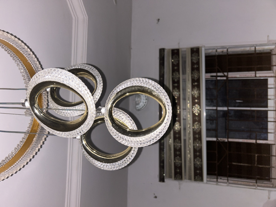 A modern chandelier with multiple gold-rimmed crystal rings hangs against a light gray wall. A window with a dark frame and metal security bars is draped with a patterned curtain, partially obscuring the view to the outside. The room appears to be dimly lit.