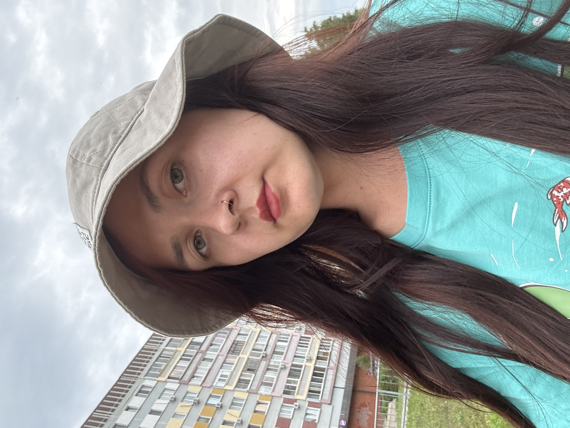 A young woman with long, dark brown hair and a gray bucket hat poses for a selfie outdoors. She is wearing a turquoise t-shirt with a graphic design. She has a nose ring and subtle makeup. In the background, a multi-story residential building with colorful balconies and windows is visible, characteristic of Soviet-era architecture found in cities like Naberezhnye Chelny, Russia. The sky is overcast.