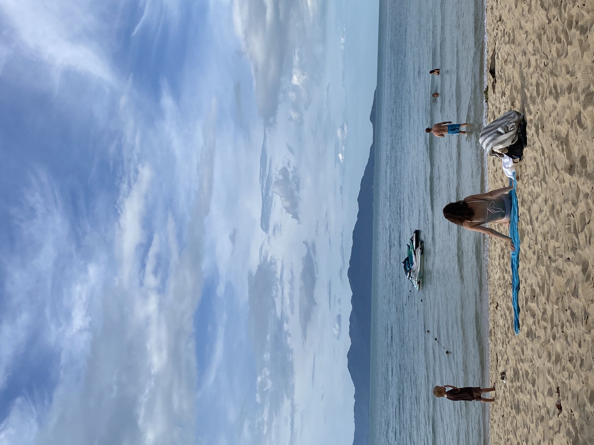 The image displays a beach scene in Baan Mae Nam, Thailand, on a partly cloudy day. In the foreground, light brown sand occupies the lower third of the frame. A person, appearing to be a woman with dark hair, is seated on the sand with her back to the camera, facing the sea. She is wearing a light-colored swimsuit and sits on a blue towel. To her immediate right on the sand, a striped towel is draped over a dark bag, beside a pair of dark shoes. Further to the left, a young, light-skinned child in a dark brown t-shirt and shorts stands facing the sea, with a hand near his mouth. A pair of sandals are visible near the child's feet on the sand.
The midground features the calm, grayish-blue ocean. A man with light skin, wearing blue swim trunks, stands partially submerged in the water, facing away from the camera. To his right and further out, two individuals are partially visible in the water. A line of dark, spherical buoys extends into the water from the left, leading towards two moored jet skis positioned further offshore. One jet ski is predominantly white and blue, while the other appears to be teal or green.
The background consists of a prominent range of dark, forested mountains across the water. The sky, occupying the upper half of the image, is covered with a mix of white and gray stratocumulus clouds, interspersed with patches of clear blue sky and some wispy cirrus clouds.