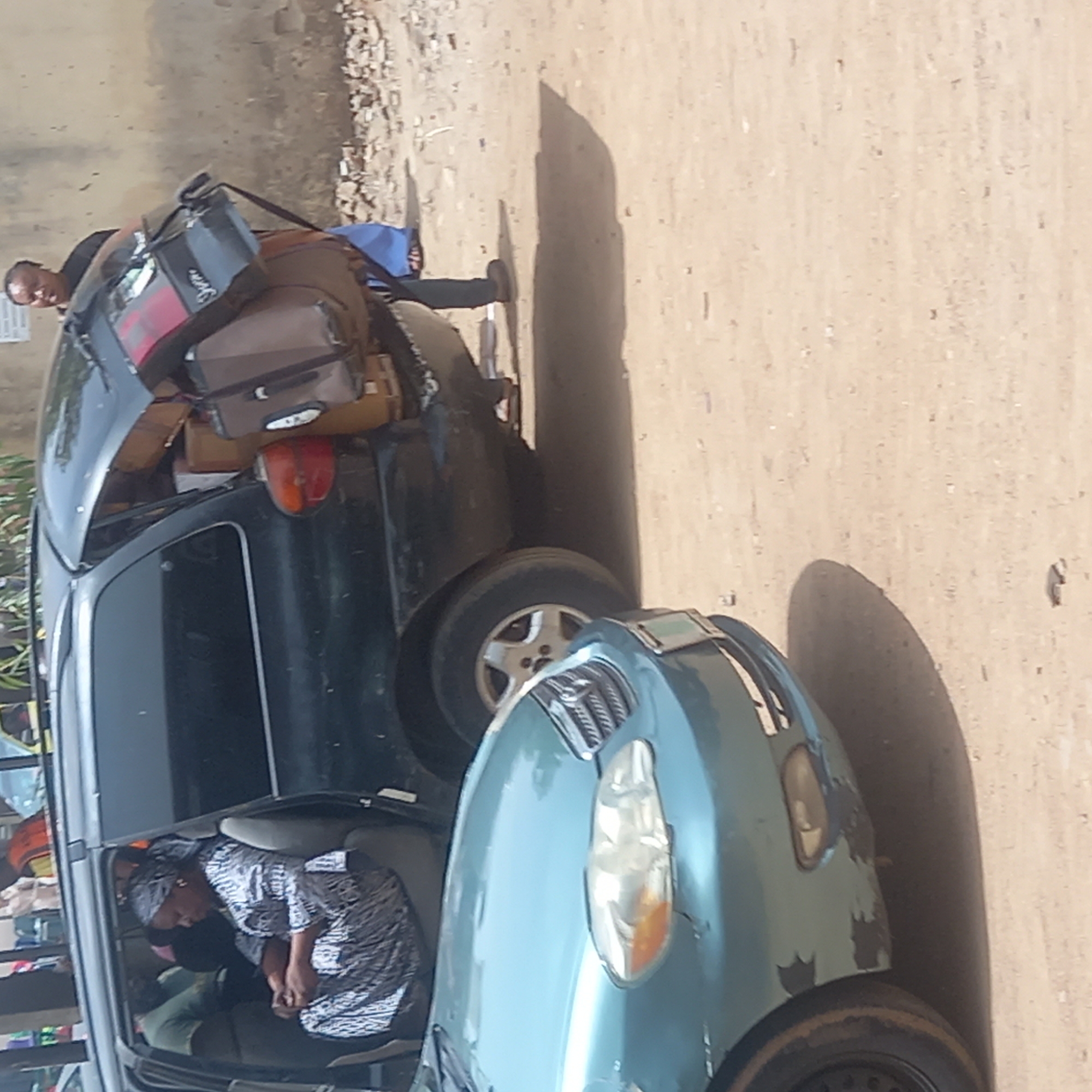 Two cars are parked side-by-side on a dusty, unpaved road in Dadi Riba, Nigeria, under bright sunlight that casts strong shadows. The car on the left is a dark-colored SUV, heavily loaded with various pieces of luggage, including brown and black suitcases and bags, strapped to its roof rack and rear. A man wearing a dark shirt stands behind the SUV, appearing to attend to or observe the loaded items. On the right, a light blue/green sedan is parked; its front fender shows visible signs of wear and tear. Inside the passenger seat of this sedan, a woman wearing a patterned top and head covering is seen leaning forward. The scene depicts a moment of travel preparation or arrival, with people and their belongings. Faint white text, possibly "Swan," is visible on one of the black bags on the SUV's roof.
