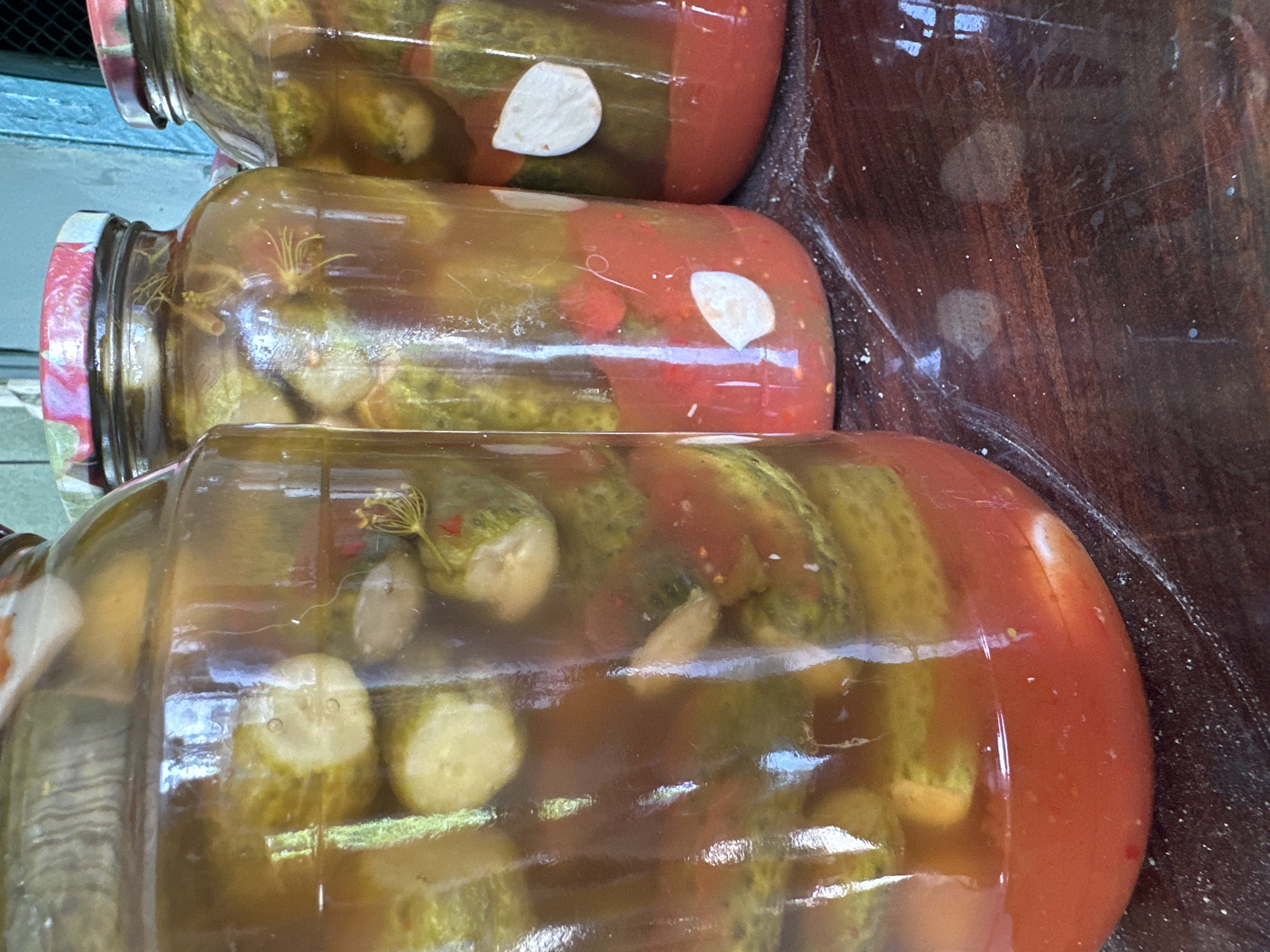 Three glass jars filled with pickled cucumbers and a red liquid are arranged on a dark wooden surface. The jars have red and white patterned lids. The cucumbers are sliced and submerged in brine, with visible pieces of dill, garlic, and possibly chili peppers. The surface they rest on is dusty and shows reflections of light. The background is blurred, but suggests an outdoor or rustic indoor setting with a light blue wall visible. The image is a close-up, focusing on the jars and their contents. There are no people or animals present in the image. The location in Yessentuki, Russia is not discernible from the visual information provided in the image.