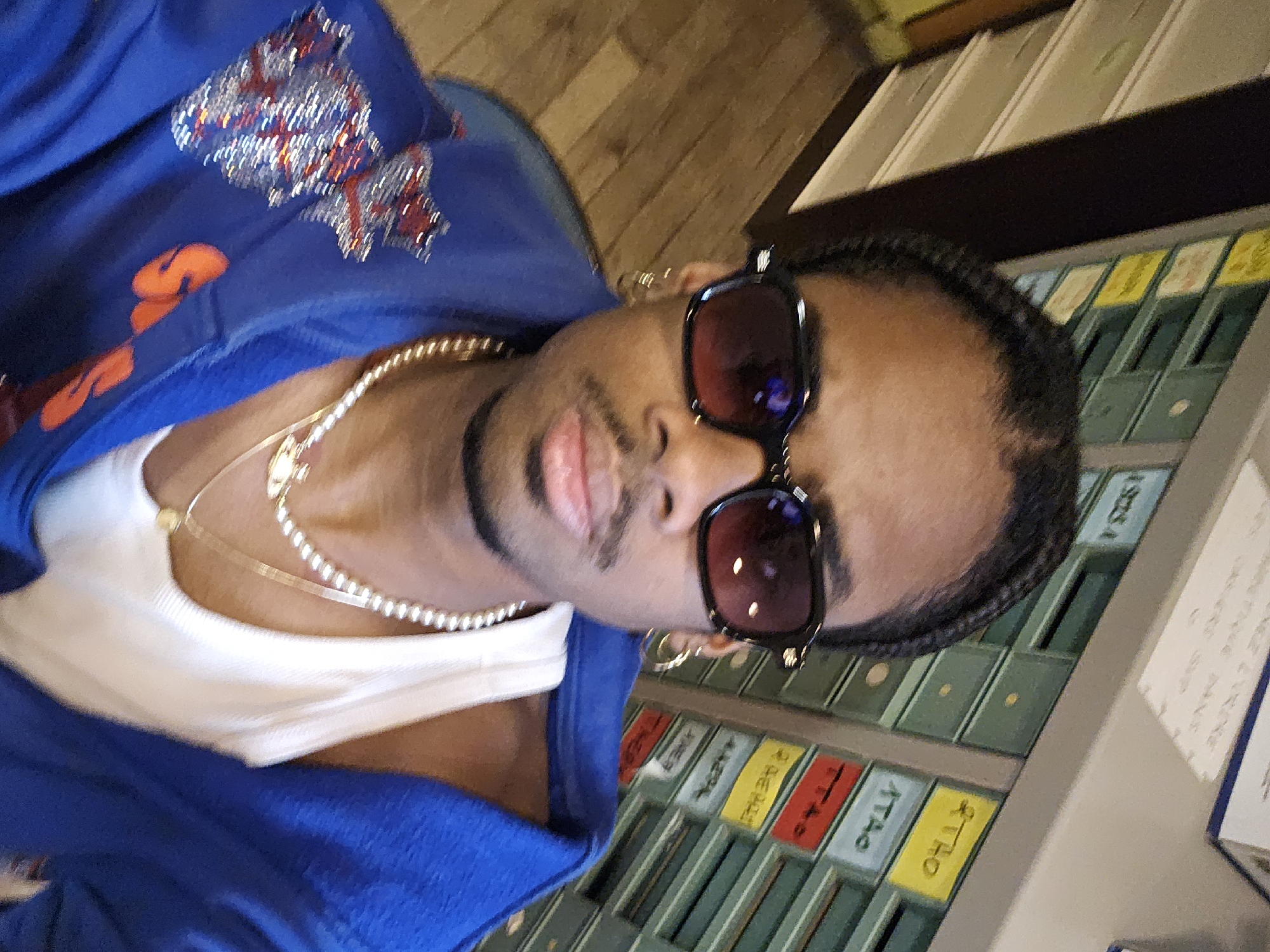 A man wearing sunglasses, jewelry, a blue jacket, and a white shirt is in a room with a wall covered in small, multicolored drawers or compartments. The drawers appear to have labels in a non-English language. The man is looking directly at the camera. The location is Paris, France.