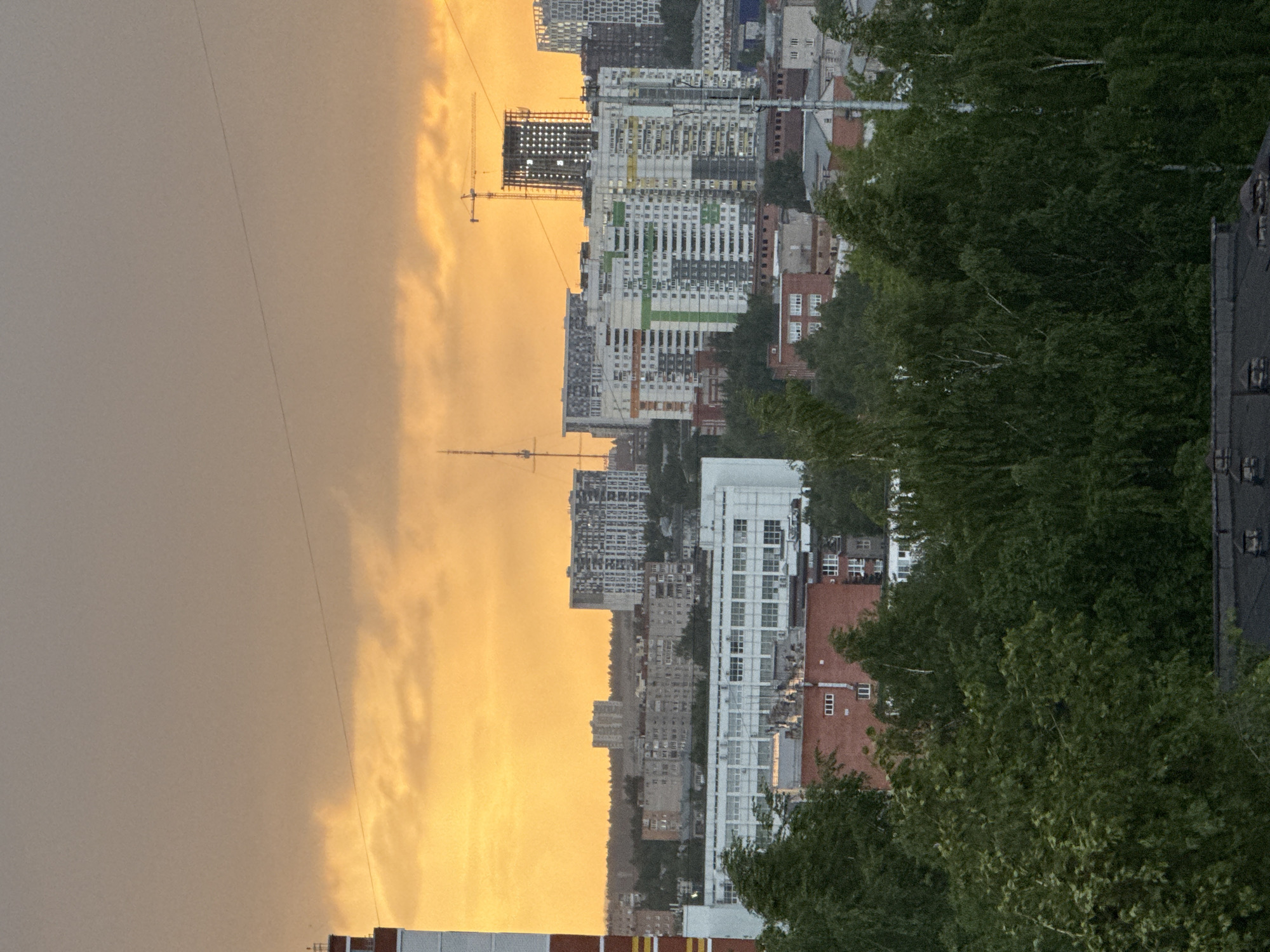 The image presents a cityscape at sunset. The photograph is vertically oriented. High-rise residential buildings dominate the central section, set against a backdrop of a golden-orange sky. Construction cranes are visible amongst the buildings. Trees are on the right side of the image, obscuring parts of the buildings. The location is Izhevsk, Russia.