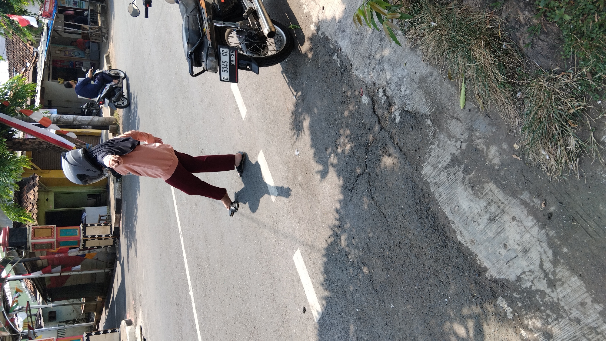 The image shows a street scene in Proyonangg, Indonesia, on a sunny day. In the foreground, a person wearing a helmet, a light pink sweatshirt, and dark red pants is walking or posing in the middle of the road, casting a shadow. The person is facing away from the camera and appears to be pointing with their right hand. A motorcycle is parked on the right side of the road, with its license plate visible. The road has white lane markings and appears to be made of asphalt. The background shows buildings, trees, and more motorcycles. There are also Indonesian flags and decorations visible, suggesting a festive or commemorative occasion. The overall environment is urban or semi-urban, with signs of local life and activity.