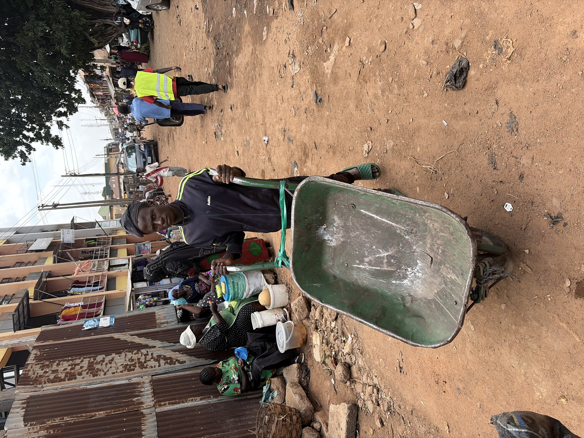 This image depicts a bustling street scene in Jos, Nigeria, during what appears to be daytime under an overcast sky. The foreground is dominated by a young man standing with a green, well-worn wheelbarrow. He wears a black and green tracksuit with the text "Feliks" visible on the sleeve and green sandals, looking directly at the camera with a neutral expression. The ground is dry, reddish-brown earth, strewn with small rocks and scattered litter.
To the left, several women are seated on the ground, which appears to be covered with corrugated iron sheets or mats. They are surrounded by various plastic containers, buckets, and a woven basket, suggesting an informal market or street vending activity. One woman is visible in a green patterned dress.
In the midground, the street is active with pedestrians and vehicles. Two men are seen walking away from the camera; one wears a bright yellow safety vest over a red shirt, and the other is in a blue shirt, pushing a smaller cart. Cars and motorcycles are visible further down the street. The background features multi-story buildings with balconies, some showing hanging laundry, indicating a dense urban or semi-urban residential and commercial area. Utility poles and power lines are also prominent against the sky. The scene captures a typical moment of daily life and commerce in the city.