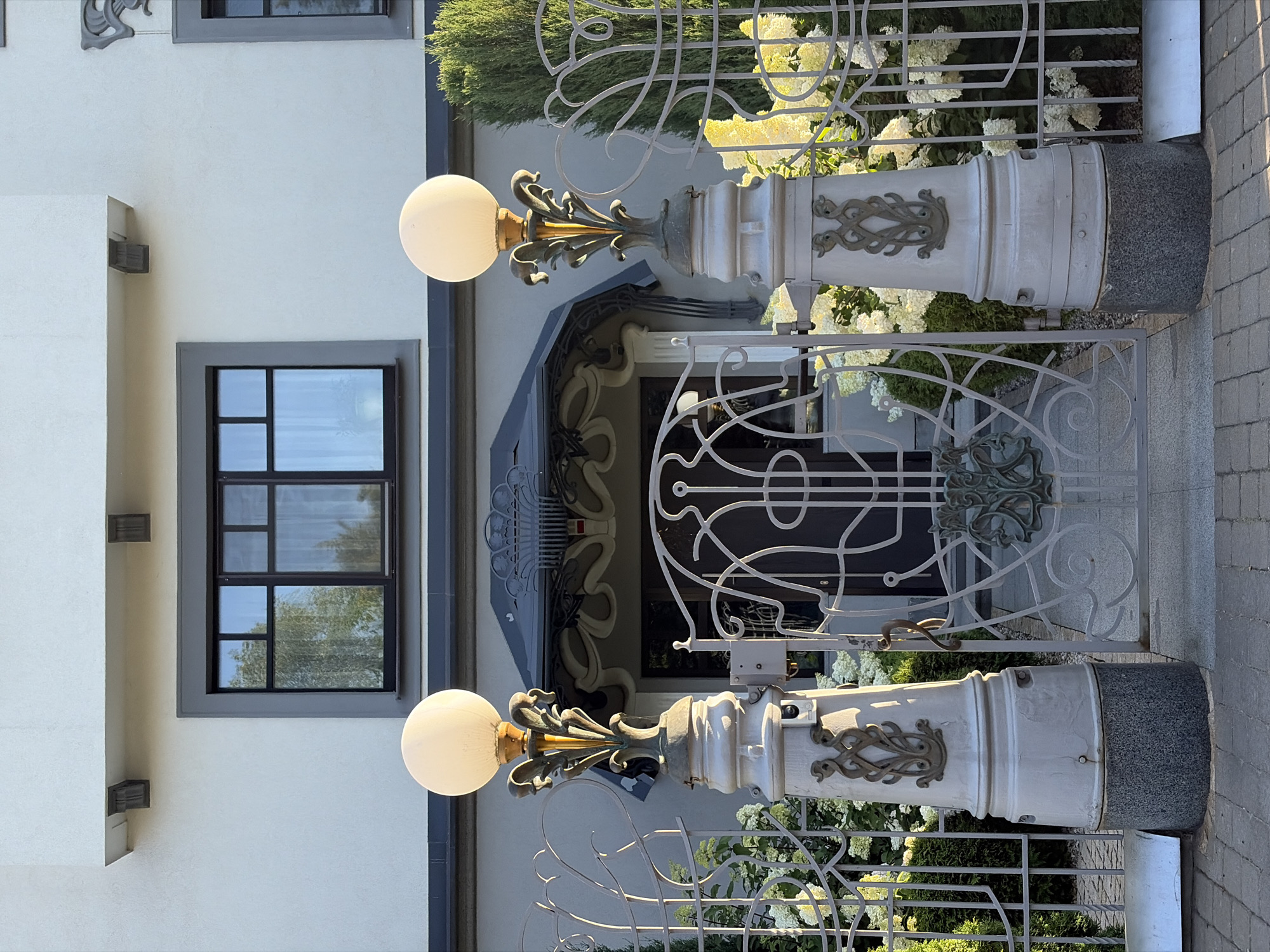 The image depicts the entrance to a building, likely a residential or public establishment, characterized by ornate architectural details. The facade is light gray, with a dark gray frame around a horizontal window with multiple panes. Above the window, two dark, wooden decorative elements protrude from the wall.
In the foreground, there is a wrought-iron gate with intricate scrollwork. The gate is flanked by two large, decorative lampposts, each topped with a globe-shaped light fixture. The lampposts are gray and feature elaborate metallic embellishments resembling stylized leaves or tendrils. Behind the gate, the entrance to the building is visible, featuring a smaller, decorative awning with curved metalwork. Lush greenery, including white flowering bushes and a trimmed evergreen shrub, surrounds the entrance and gate, adding a touch of natural beauty to the scene. The ground is paved with cobblestones.
This scene is located at the Central Department Store (TsUM) in Minsk, Belarus. The architectural style suggests a historical or art nouveau influence, which is consistent with some of the older buildings in the Belarusian capital. No people are present in the image. The lighting suggests it is daytime, likely late afternoon due to the warm cast of sunlight on the lampposts and foliage.