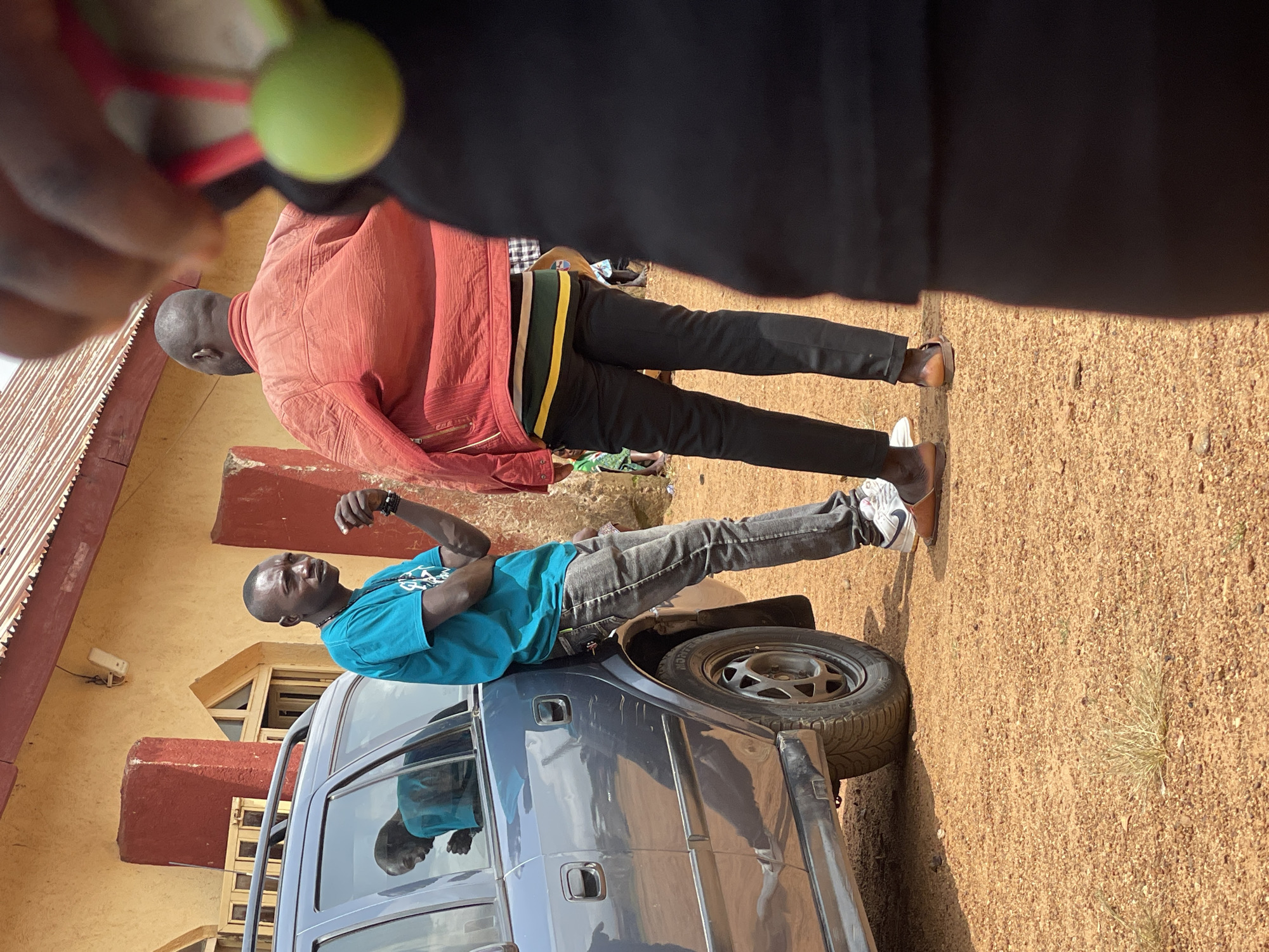 In Kassa, Nigeria, a man in a teal t-shirt leans against a dark grey car. He has his left arm bent, with his hand near his chin, and his right arm resting on the car. He wears grey jeans and white sneakers with a Nike logo. Another man, wearing a red jacket over a horizontally striped shirt with green, yellow, and white stripes, and black pants, stands facing away from the camera, talking to the first man. His left foot is visible, wearing brown sandals. The ground is covered in reddish-brown gravel, with dry grass patches. Behind the men is a building with a yellow facade and a red, corrugated metal roof. The sky is overcast. A blurry object, possibly a person holding a green object, is in the foreground on the right.