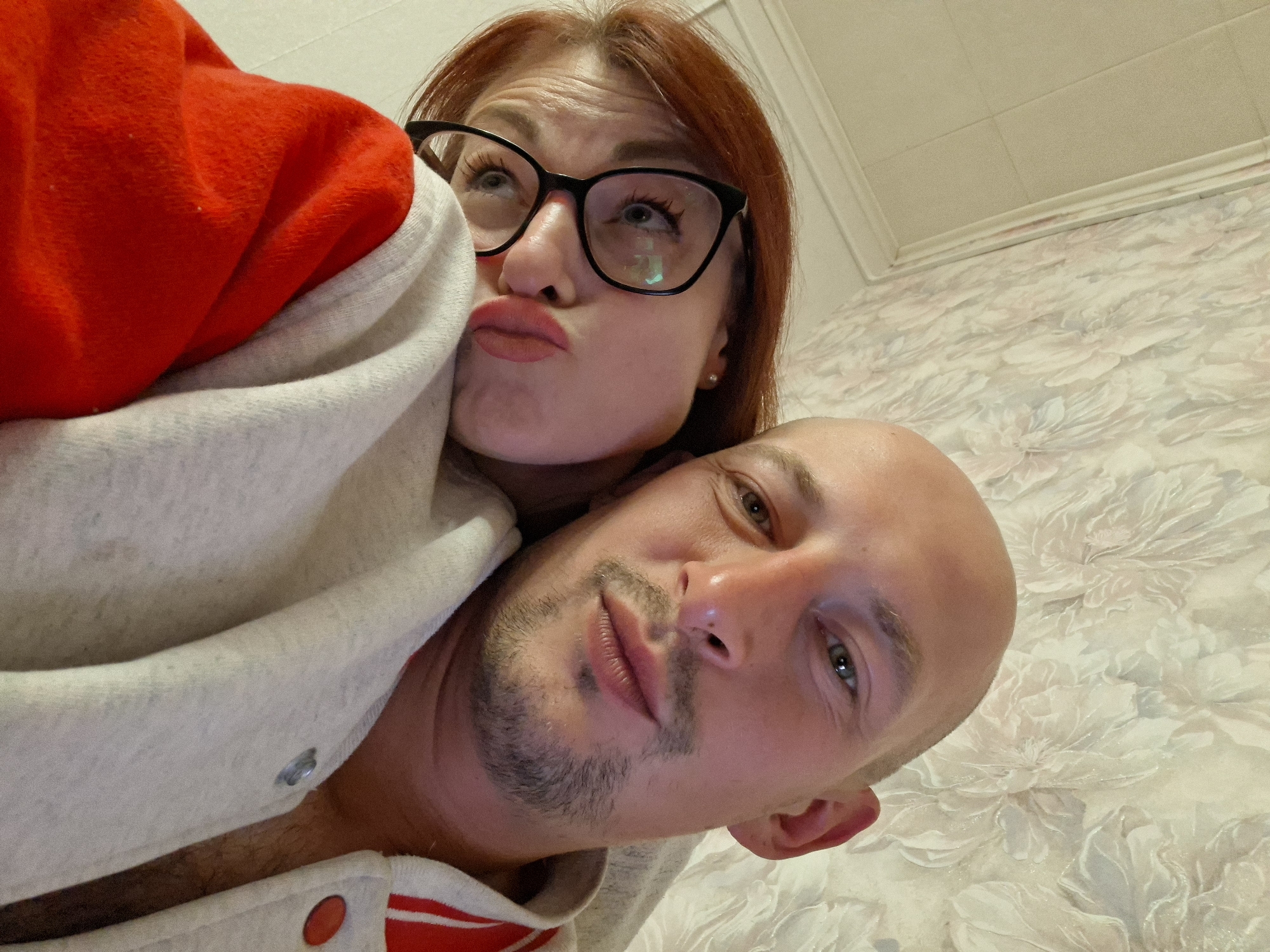 In a close-up, low-angle selfie, a man and a woman are positioned in the foreground. The man's head is angled upwards, with his face and the top of his bald head filling the right side of the frame. He has green eyes and a light beard and mustache. The woman is nestled behind him, her face tilted downwards and her lips pursed. She has red hair and is wearing black-rimmed glasses. They are both wearing casual clothing, with a red garment visible on the woman and a light-colored hooded jacket on both. The background consists of a textured wallpaper with a floral pattern and a white ceiling. The scene appears to be indoors, possibly in a residential setting. There is no explicit indication of the specific location within Забалоцкі сельскі Савет, Belarus.