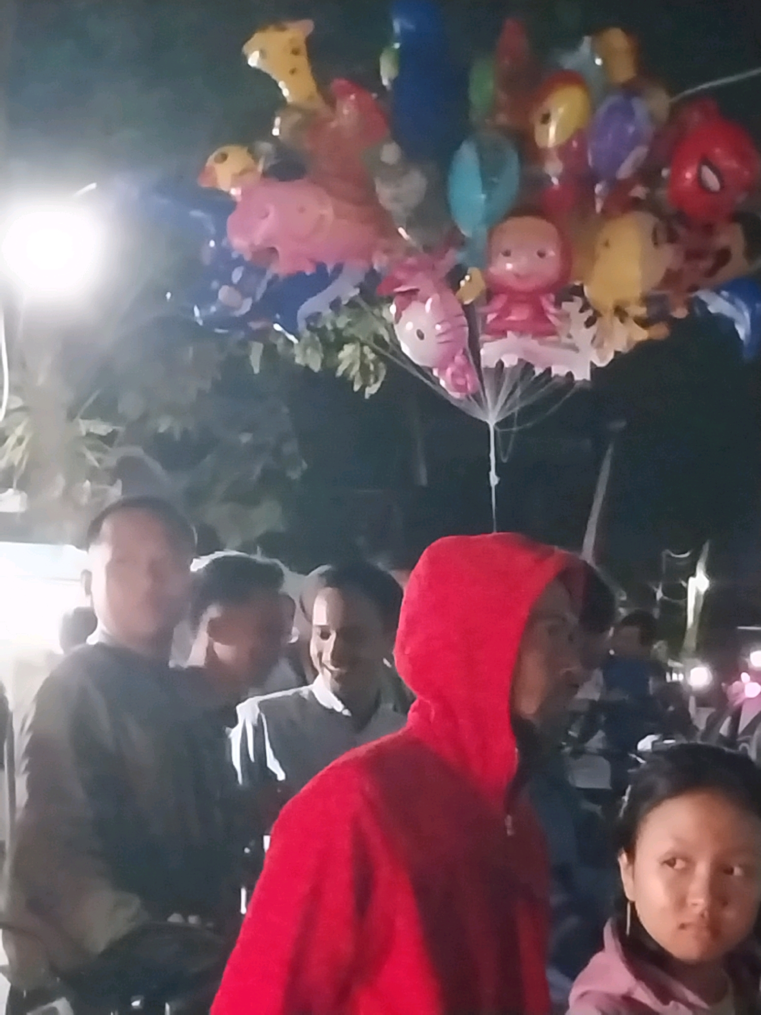 An outdoor, nighttime scene in what appears to be a public gathering or market area in Nglegok, Indonesia. The image is blurry and low-resolution, making precise identification difficult. In the foreground, a person wearing a red hooded sweatshirt is partially visible from the shoulders up, facing to the right. To their right, a young female with dark hair and a light-colored jacket is also looking to the right. Behind them, several other individuals are present, their faces and attire indistinct due to the low quality and lighting. In the upper portion of the image, a cluster of colorful balloons is visible, some appearing to be character-shaped, such as Hello Kitty and Spider-Man. The background is dark with some faint lights suggesting artificial illumination. The overall impression is of a lively, possibly festive, outdoor event.
