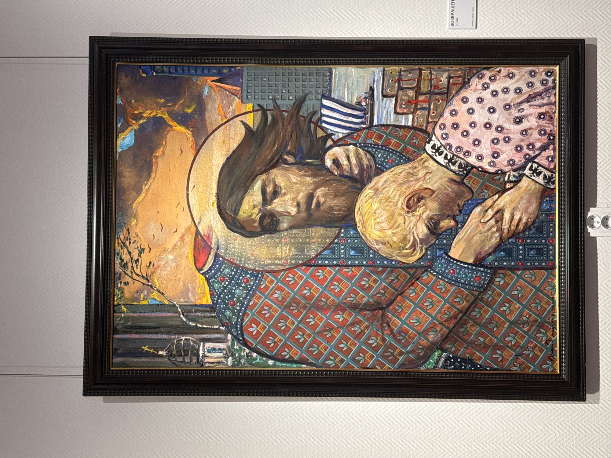 The image displays a framed oil painting titled "Возвращение" (Return) from 2004. The painting depicts two figures, one larger and one smaller, against a backdrop that blends religious and urban elements. The larger figure, with a halo, has a flowing beard and hair, and wears a richly patterned garment in red and green with diamond shapes. The smaller figure, with blond hair, is bowing down, kissing the hand of the larger figure, and is dressed in a pink garment adorned with circular patterns.
The background features architectural details such as a church dome with a cross on the left, a barren tree branch with flying birds in the sky, and modern buildings on the right, suggesting a city. A body of water with a boat and a ship with striped sails is also visible. The overall style is expressive and symbolic, with bold colors and textures. A small white label in the lower right corner of the painting indicates the title and year.
The painting is displayed on a textured white wall, typical of a gallery setting. The lighting appears to be even, highlighting the artwork. Below the painting, a small label with Cyrillic text and what appears to be an artist's signature is visible. There are also mounting fixtures for the painting visible on the wall. This scene is not in Novosibirsk, Russia, but rather in a gallery setting where the artwork is displayed.