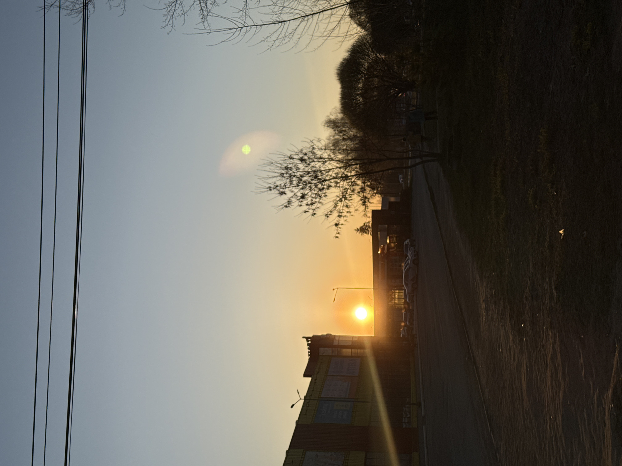 The image shows a sunset scene in an urban environment, possibly in the city of Unknown, Unknown. The sun is setting behind a building, creating a bright glare and rays of light. A street runs alongside the building, with cars parked along the side. Trees and power lines are visible in the foreground and background. The sky transitions from a light blue to a yellow-orange gradient. The area to the right of the street appears to be an unpaved ground.