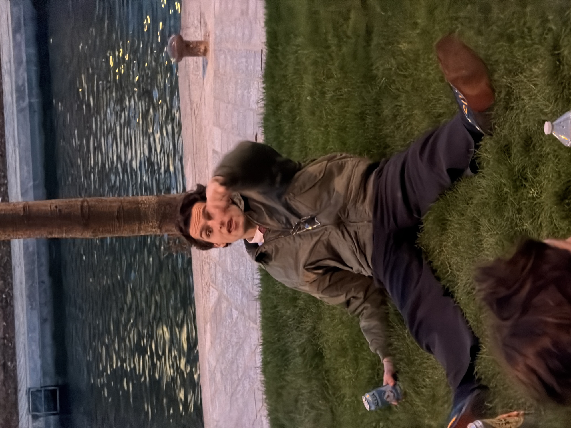 A man lies on the grass next to a body of water, possibly the Seine River. The location appears to be an area near the river in Paris, France, given the architectural details and general ambiance. The man is wearing dark-colored pants and a tan jacket, with brown shoes. He has a can in his hand. Another person is partially visible in the lower right of the image. The setting suggests a casual, possibly evening, scene along the riverbank.