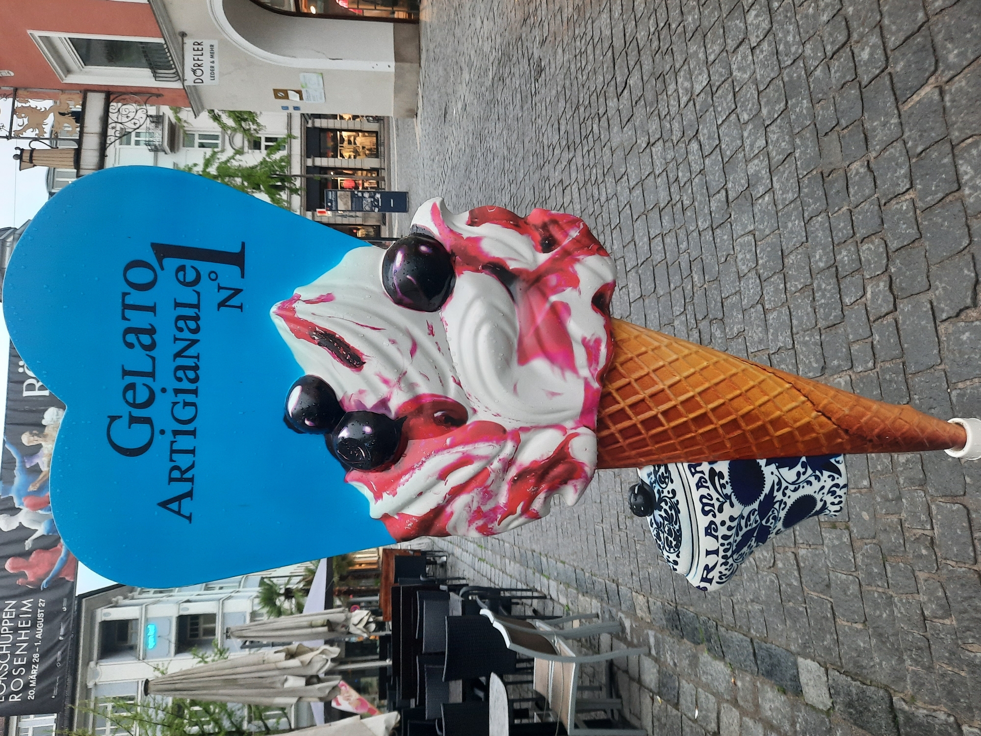 This image depicts a large, decorative ice cream cone sign placed on a cobblestone street in Rosenheim, Germany. The sign is blue and heart-shaped, with the text "GELATO ARTIGIANALE N°1" prominently displayed. The ice cream itself is depicted as white with red swirls and topped with dark berries.
In the background, there are buildings with storefronts, outdoor seating areas with umbrellas and chairs, and a banner that reads "ROSEHEİM" and "20. MÄRZ 26 – 1. AUGUST 27". A store sign reads "DÖRFLER LEDER & MEHR". The overall environment suggests a commercial area in a European city, likely a pedestrian zone given the cobblestone street and outdoor seating. No people are clearly visible in the frame.